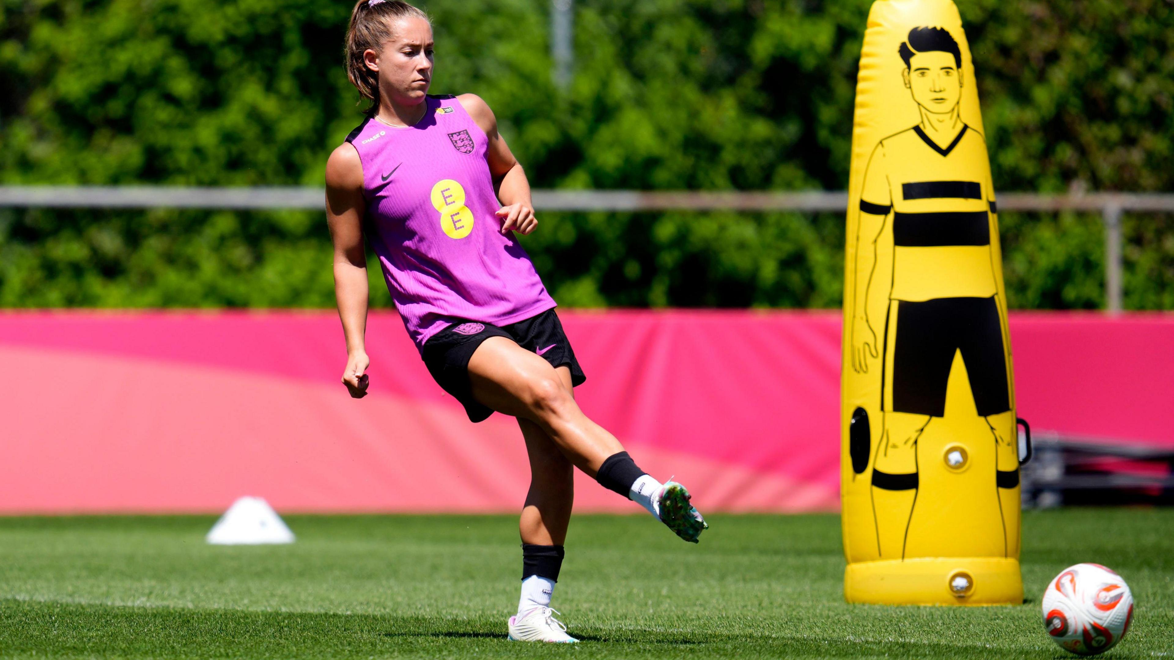 A brown-haired woman in a purple England training top passes a football on a training ground with a yellow, blow-up football dummy and some cones behind her.