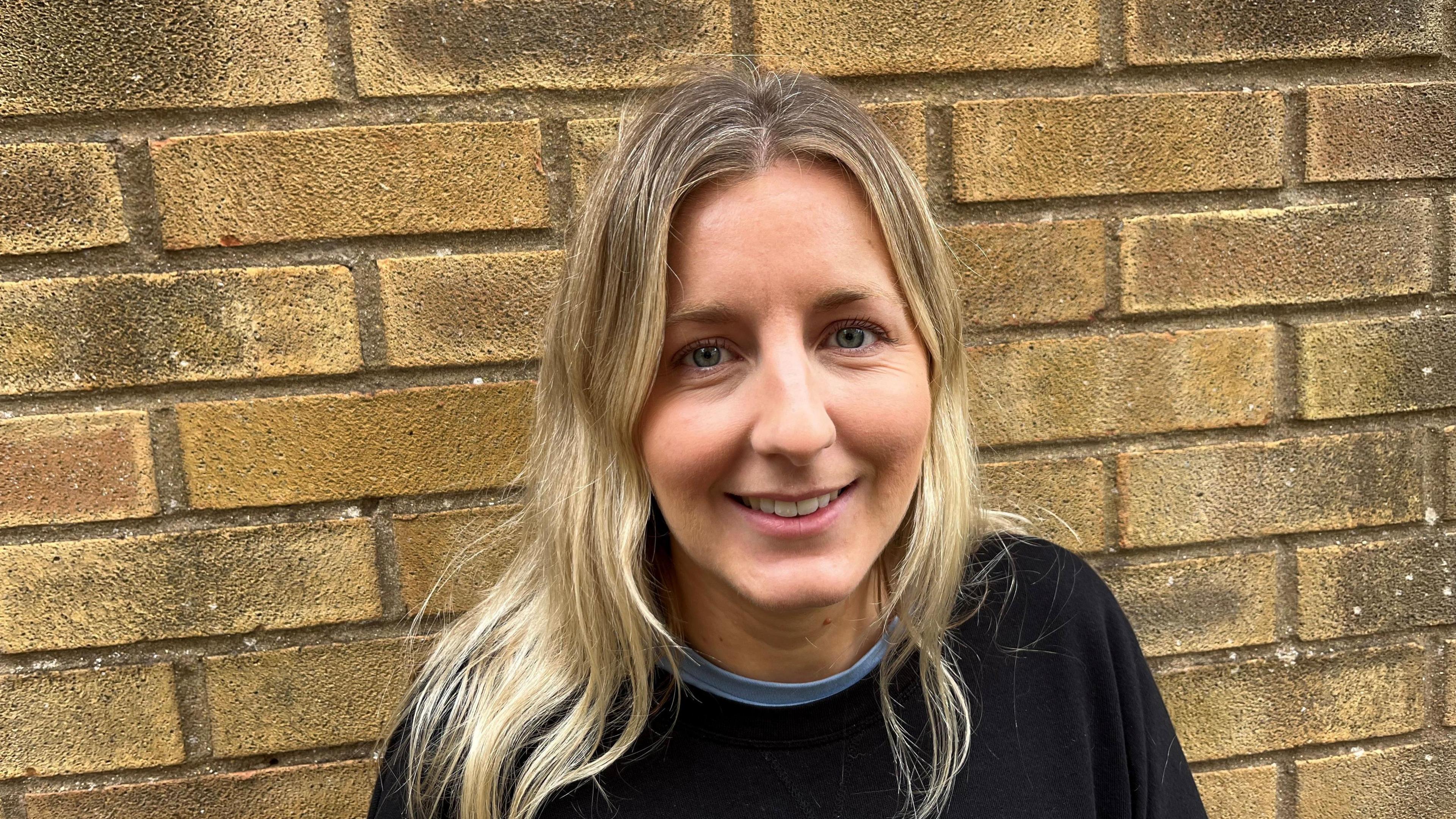 Clare Biddle, 32, has long, shoulder length blonde hair, with a centre parting, she is wearing a black jumper with a blue shirt underneath, she is standing in front of a brick wall.