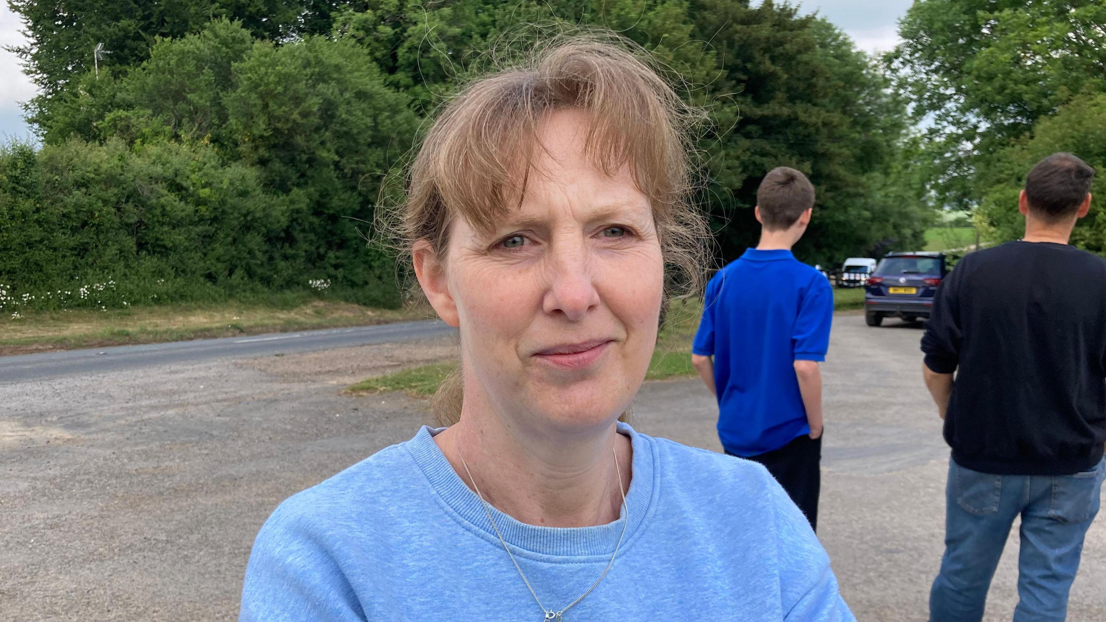 A photo showing a woman with brown hair, wearing a blue jumper. She is stood in a layby next to a country road. There are two people and a car in the background.