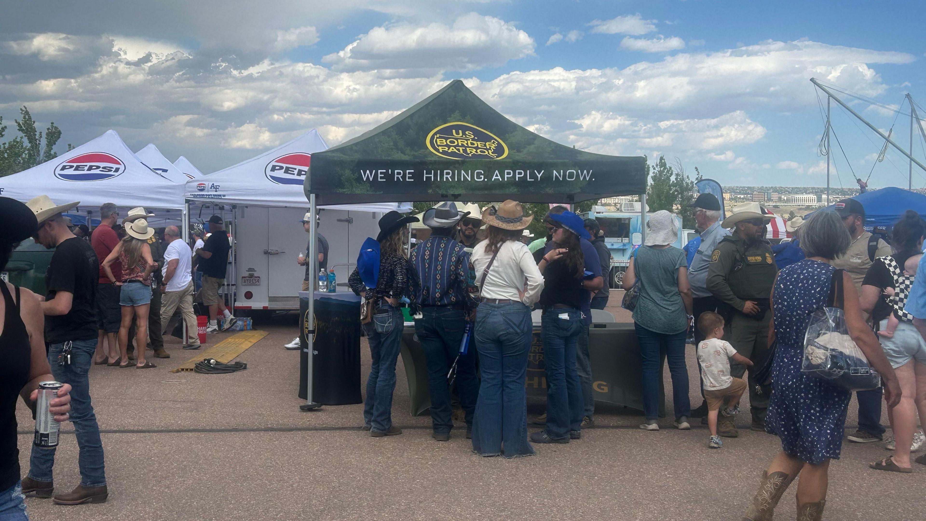 Folks wearing cowboy hats and jeans line up in front of a tent emblazoned with the Border Patrol logo that reads "We're hiring. Apply now"