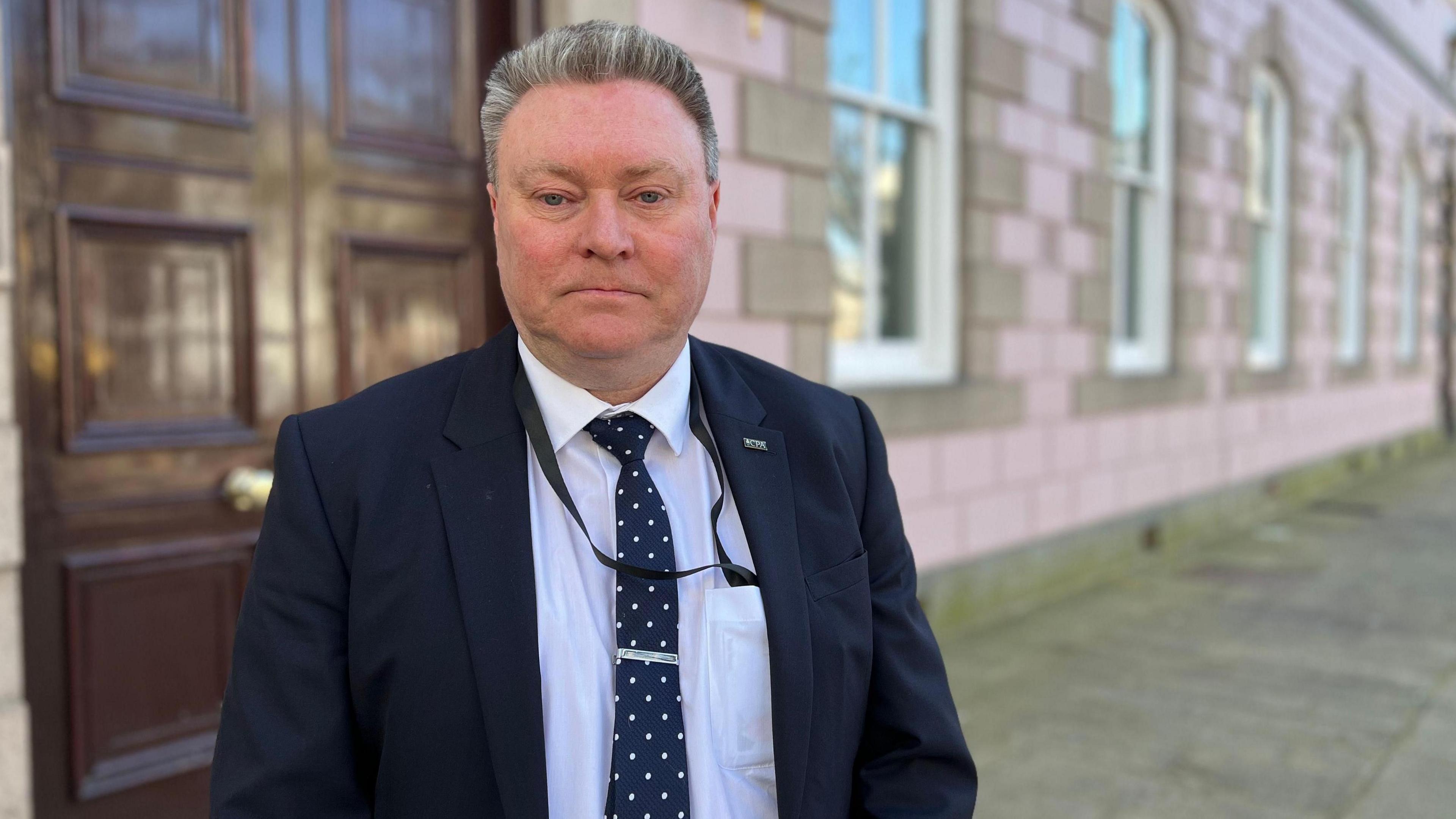 Jersey education minister Deputy Rob Ward stands outside a government building which has a large brown wooden door and lilac coloured brick work. He is wearing a dark suit and a white shirt with a black and white polka dot tie.
