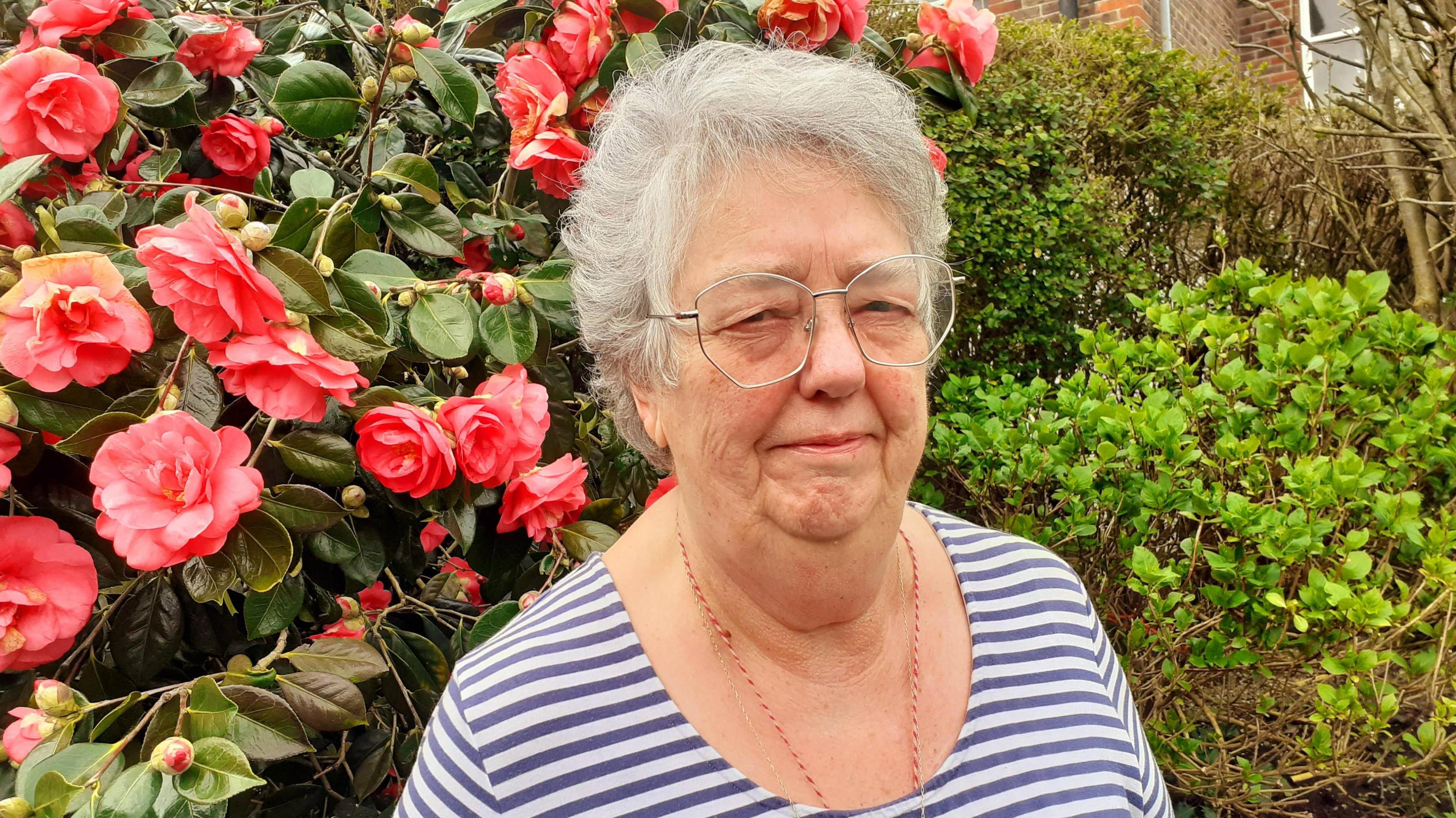 White senior woman with glasses stands in front of a pink flower bush