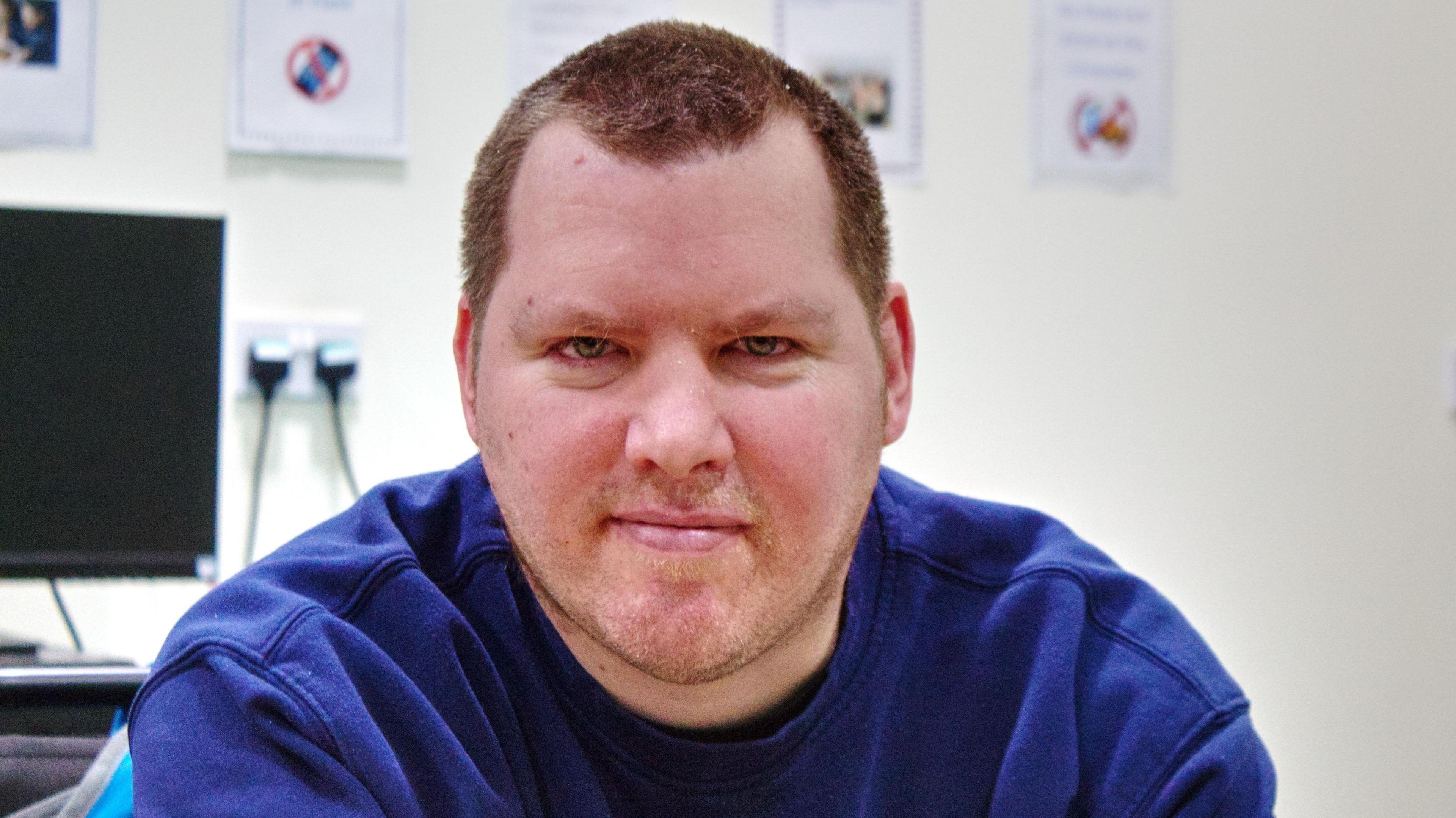 Barry, looking straight at the camera, wearing a blue top, he has short hair, and some stubble on his face. There is a computer behind him to the left. 