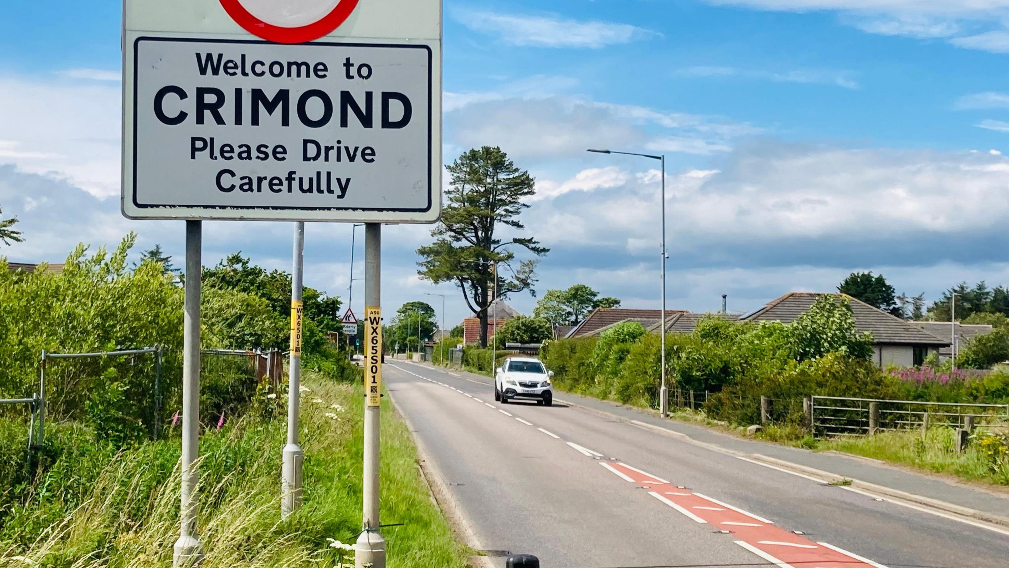 A street sign that says welcome to crimond beside a road with a lone white car under a blue sky with white clouds.