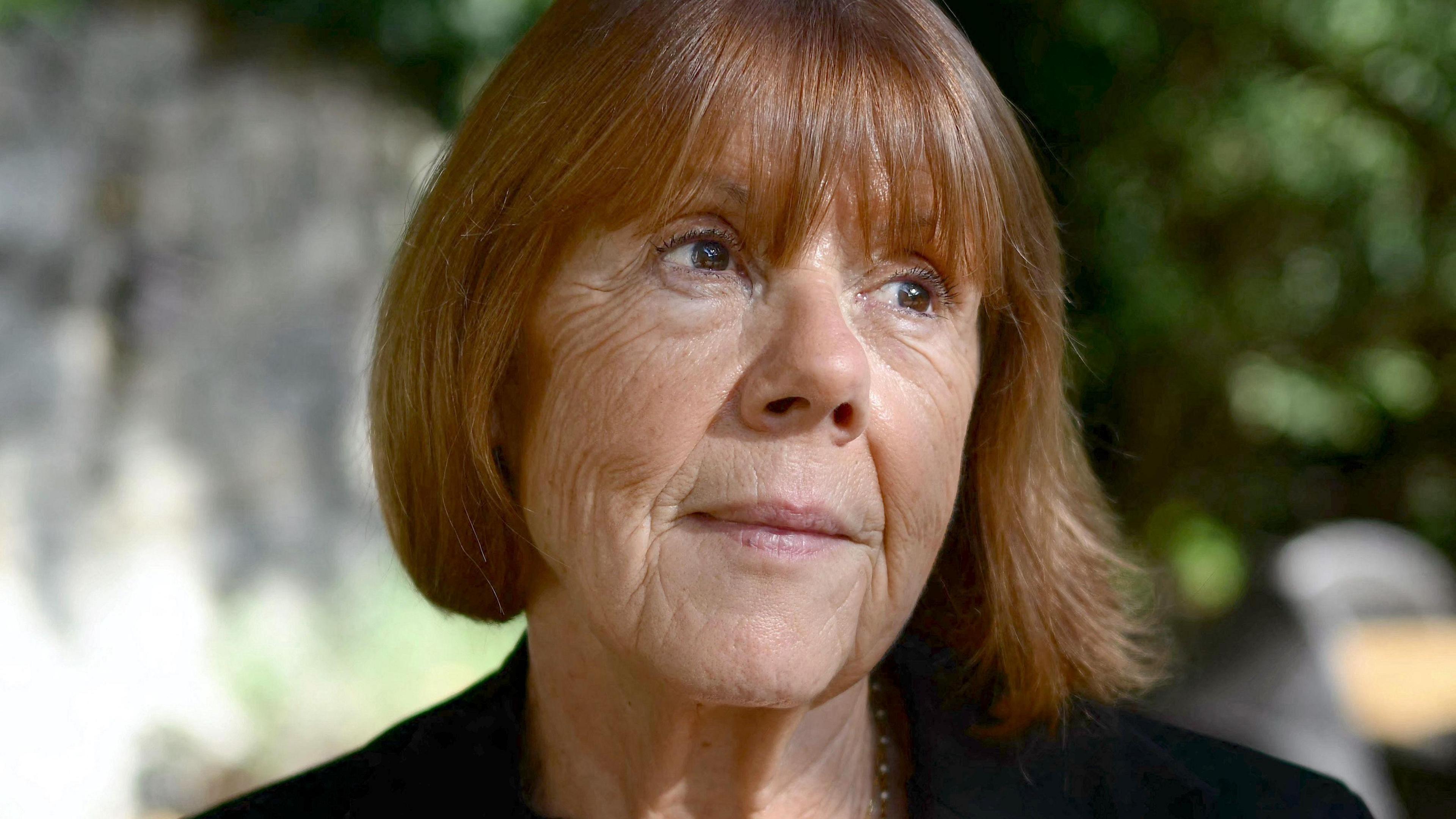 Gisèle Pelicot, a woman with chestnut hair in a bob, pictured in the dappled light of a garden or park, in close-up, looking up and to the left, with the background blurred behind her