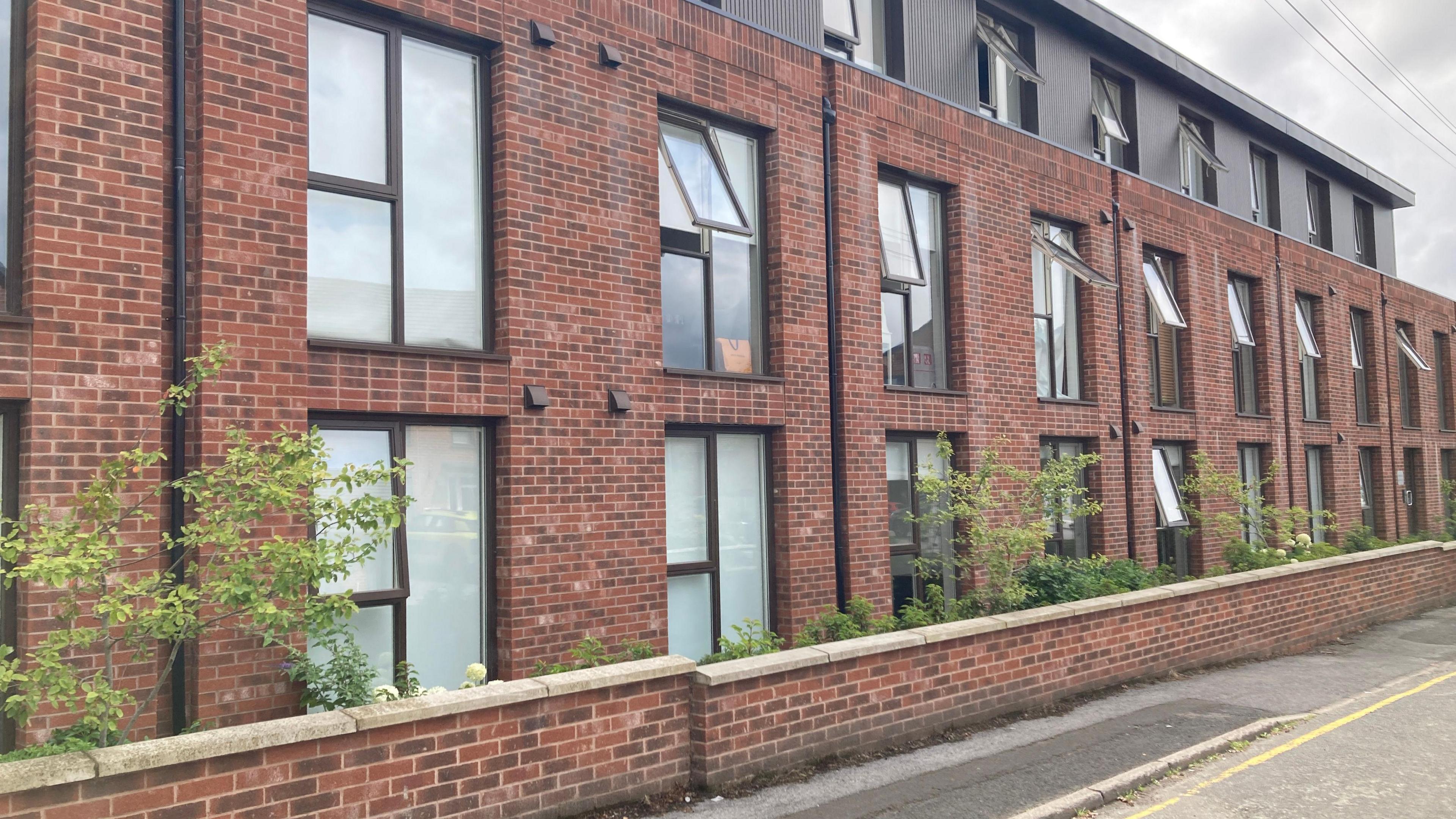A view of Valley Court in Sandbach, a modern red-brick building with a second storey in black cladding, set back from the ground and first floors. Trees and bushes are sprouting between the building and the boundary wall onto the street.