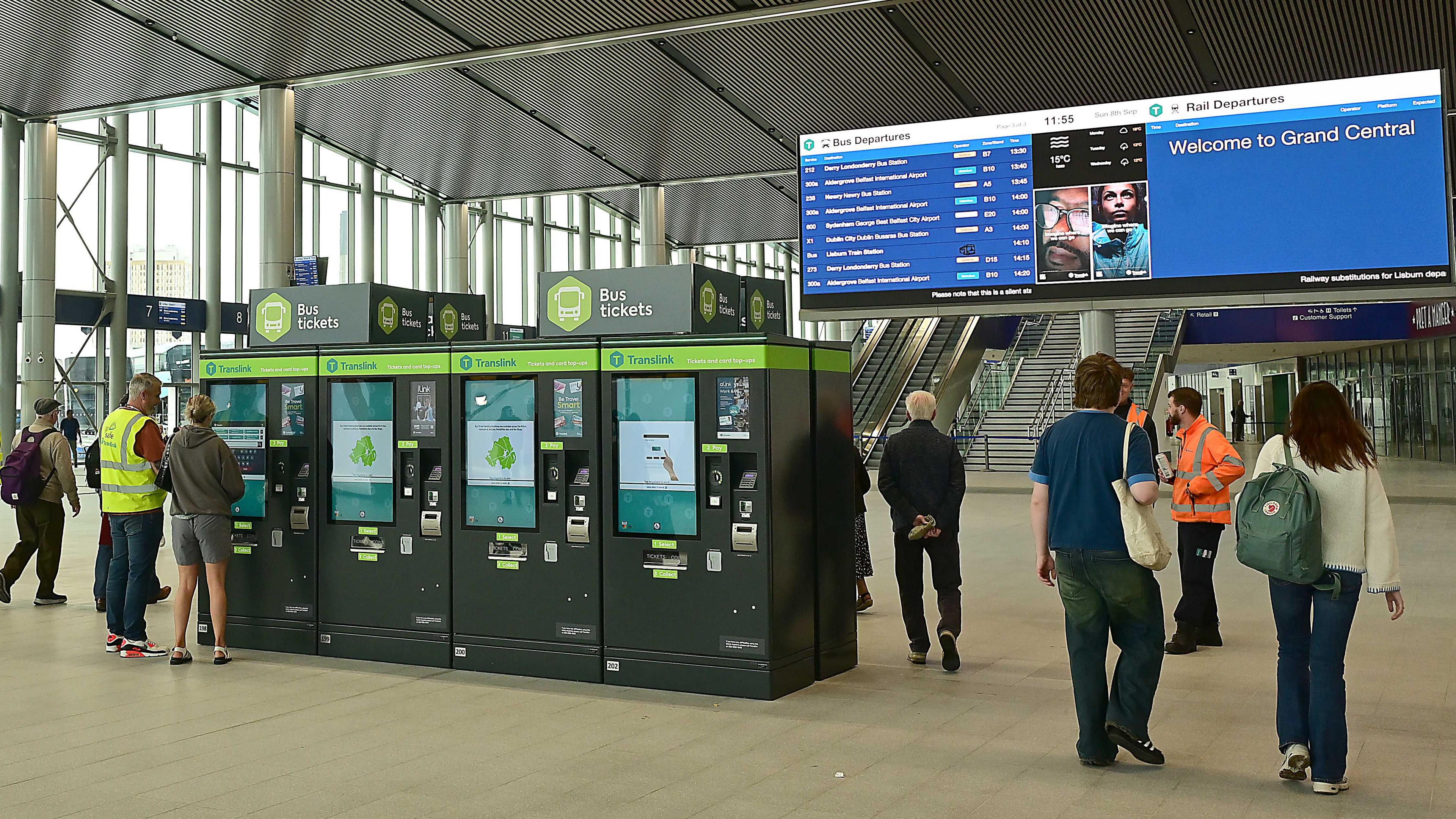 Passengers walking about Grand Central Station in Belfast. In the foreground some ticket machines. In the background a large screen with departure times on one side and "Welcome to Grand Central" on the other.