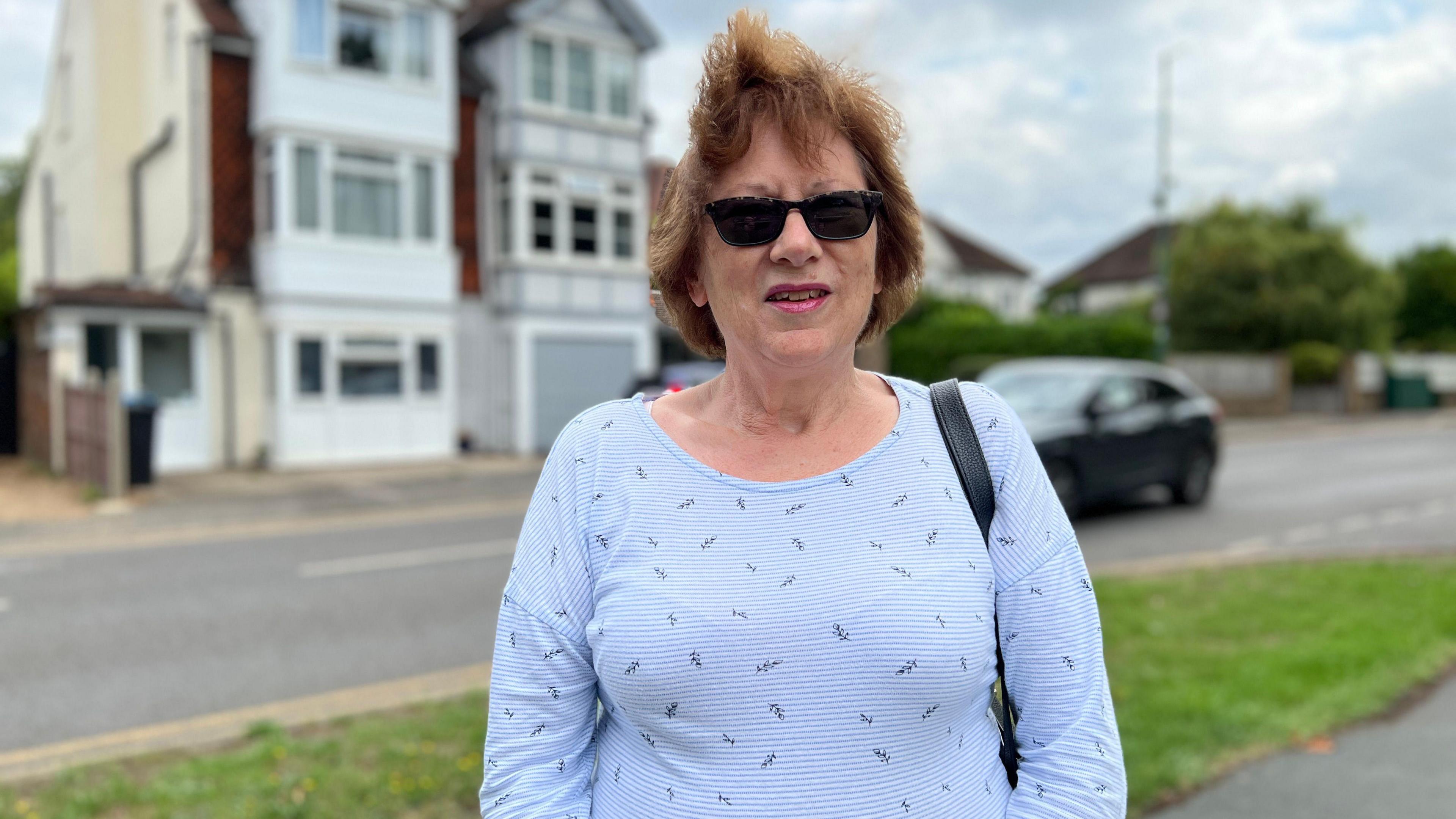 Charmian Davies has short brown hair and is wearing a blue and white stripy top and a pair of sunglasses. She's standing on a grass verge and looking straight at the camera. There's a road, black car and a house behind her.