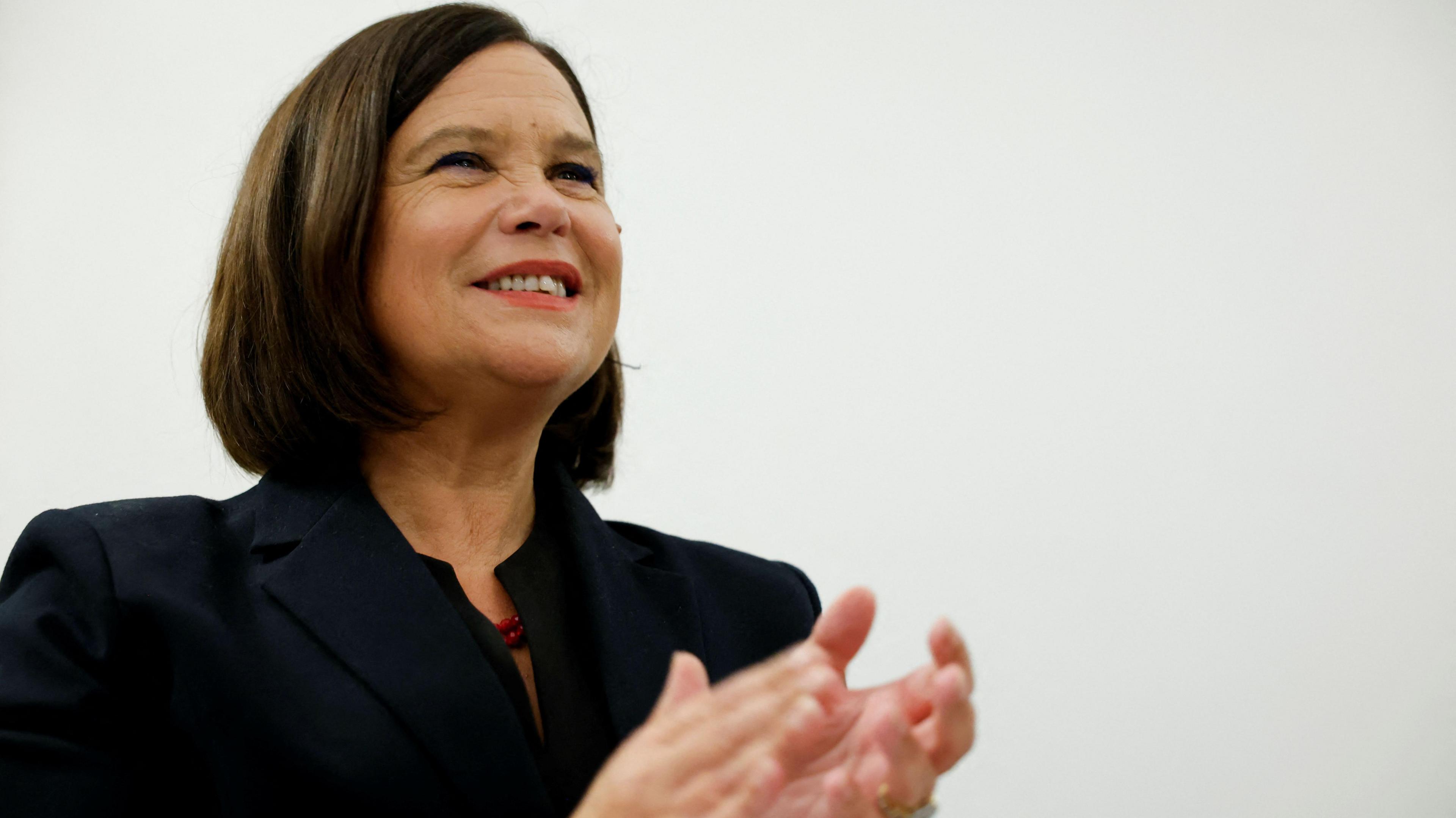 Mary Lou McDonald - a woman with short brown hair above her shoulders applauds while standing against a white background. She is wearing a dark suit jacket and a red, beaded necklace.