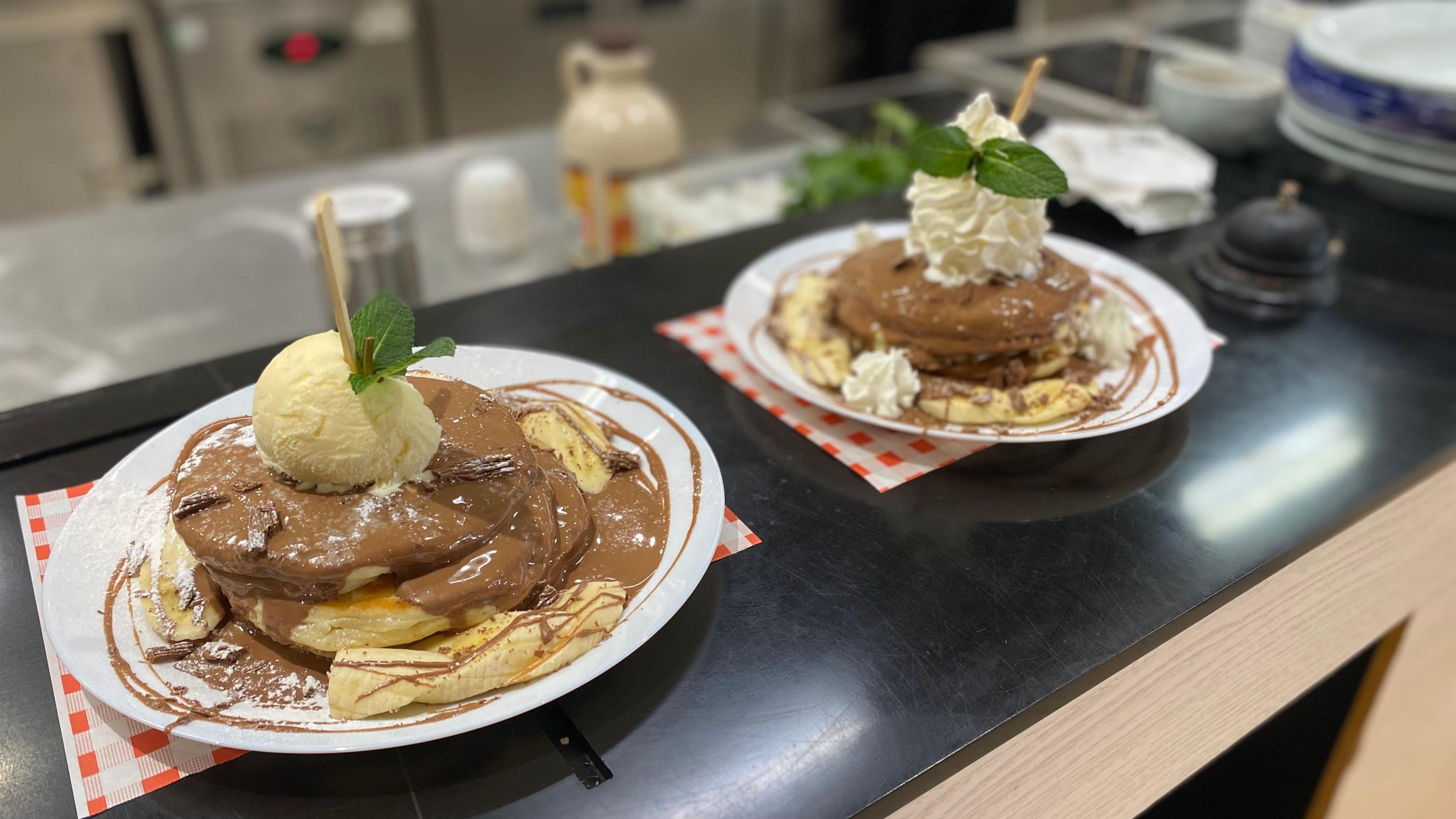 Two plates with pancakes sit on a black kitchen counter. There are two fluffy pancakes on each place, covered in chocolate sauce and whipped cream.