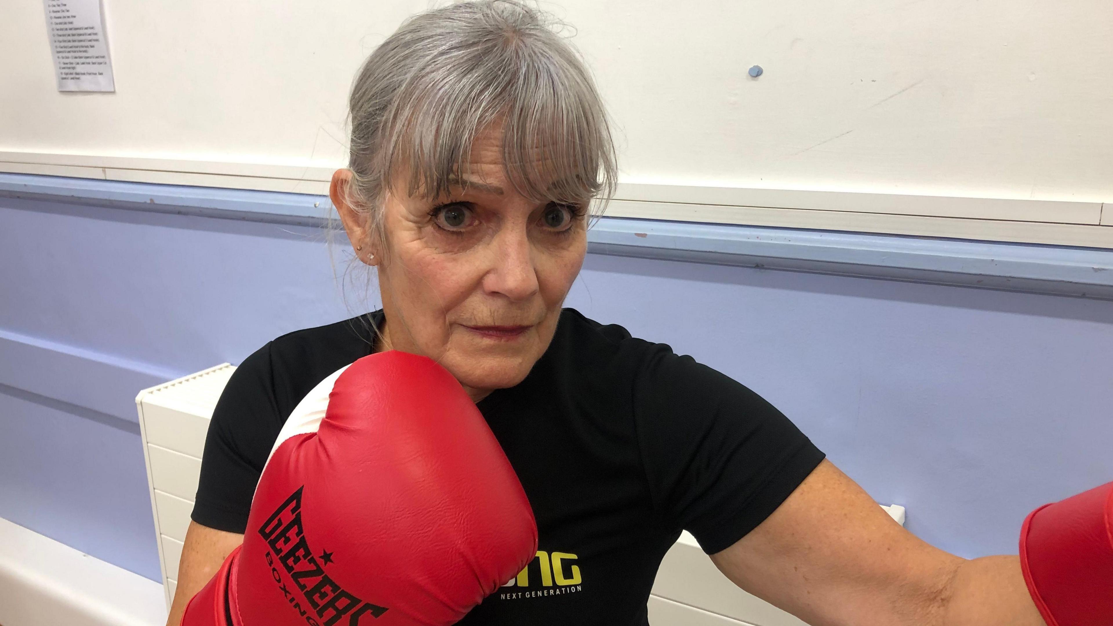 Jane Perry, 71, has grey hair over her forehead but not in her eyes. She is wearing a black t-shirt and has red and white boxing gloves on. She is in a 'boxing pose' with her right arm held near her chest and her left extended.