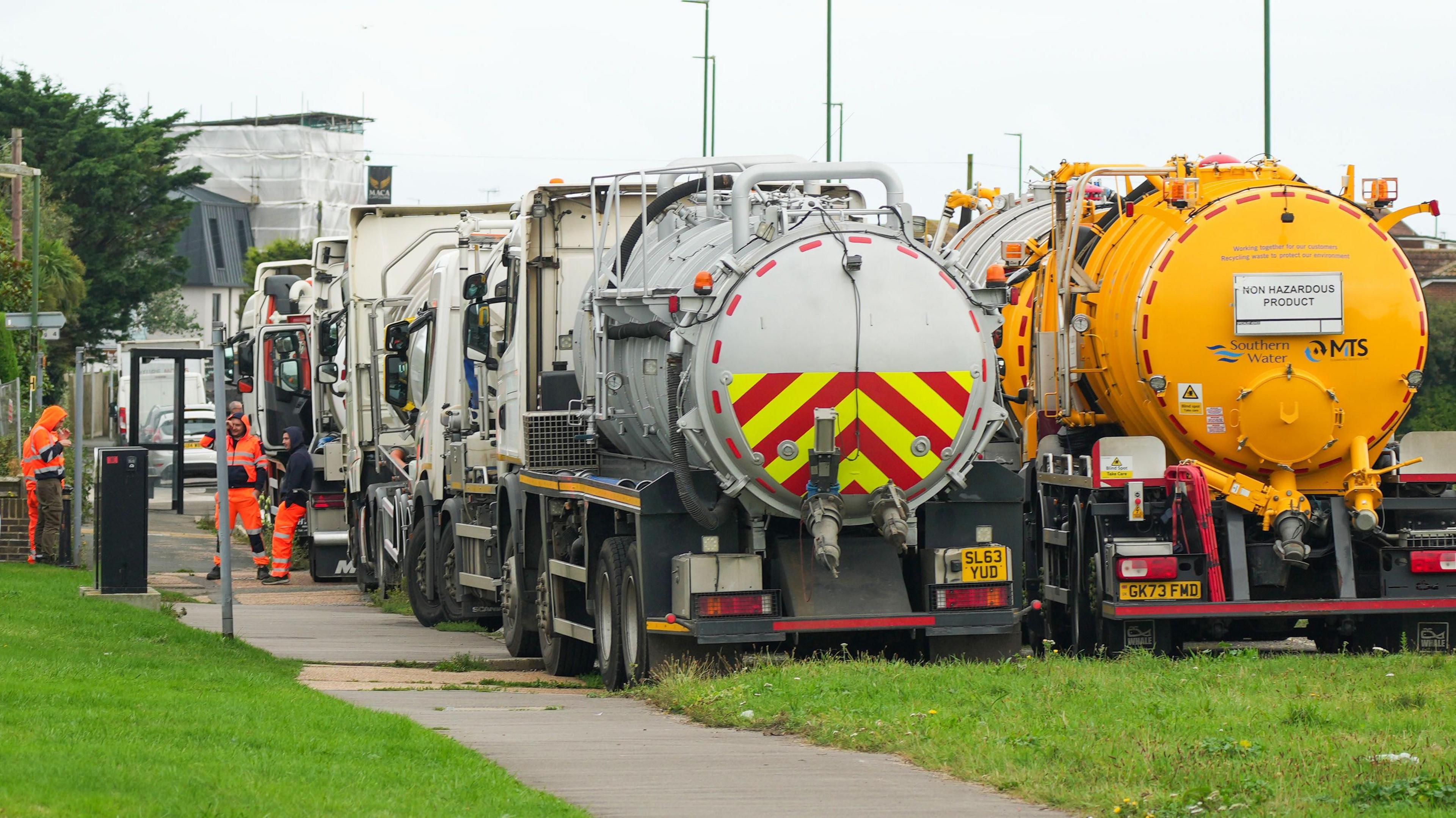 Two tankers, one white and one yellow, are parked on a grassy verge. Workers in hi-vis stand next to them chatting