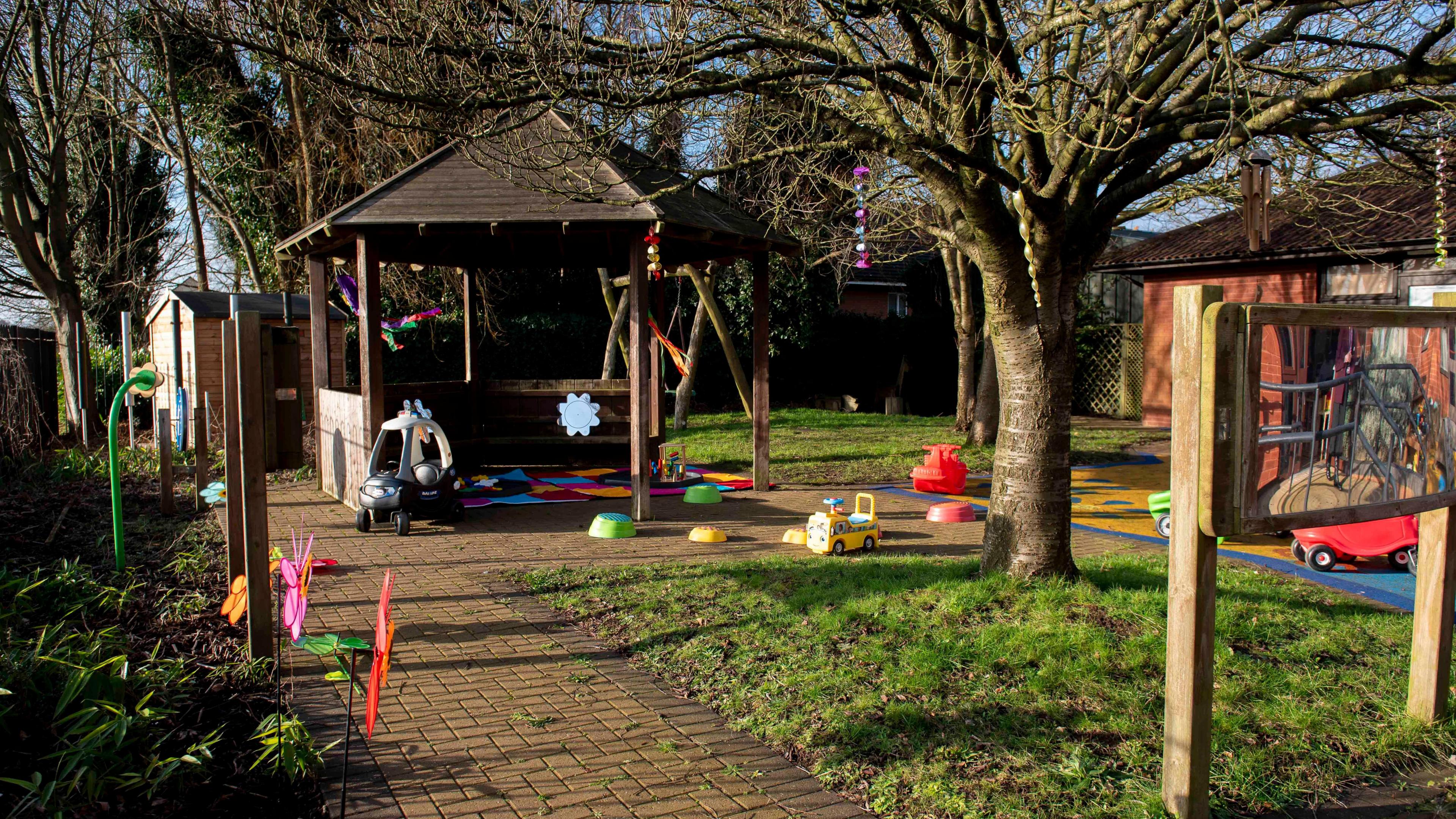 A garden with a paved path, grassy areas and trees. An outdoor seating area with pitched roof is at the end of the path and small children's toys lie around the garden including a ride-on truck and car.