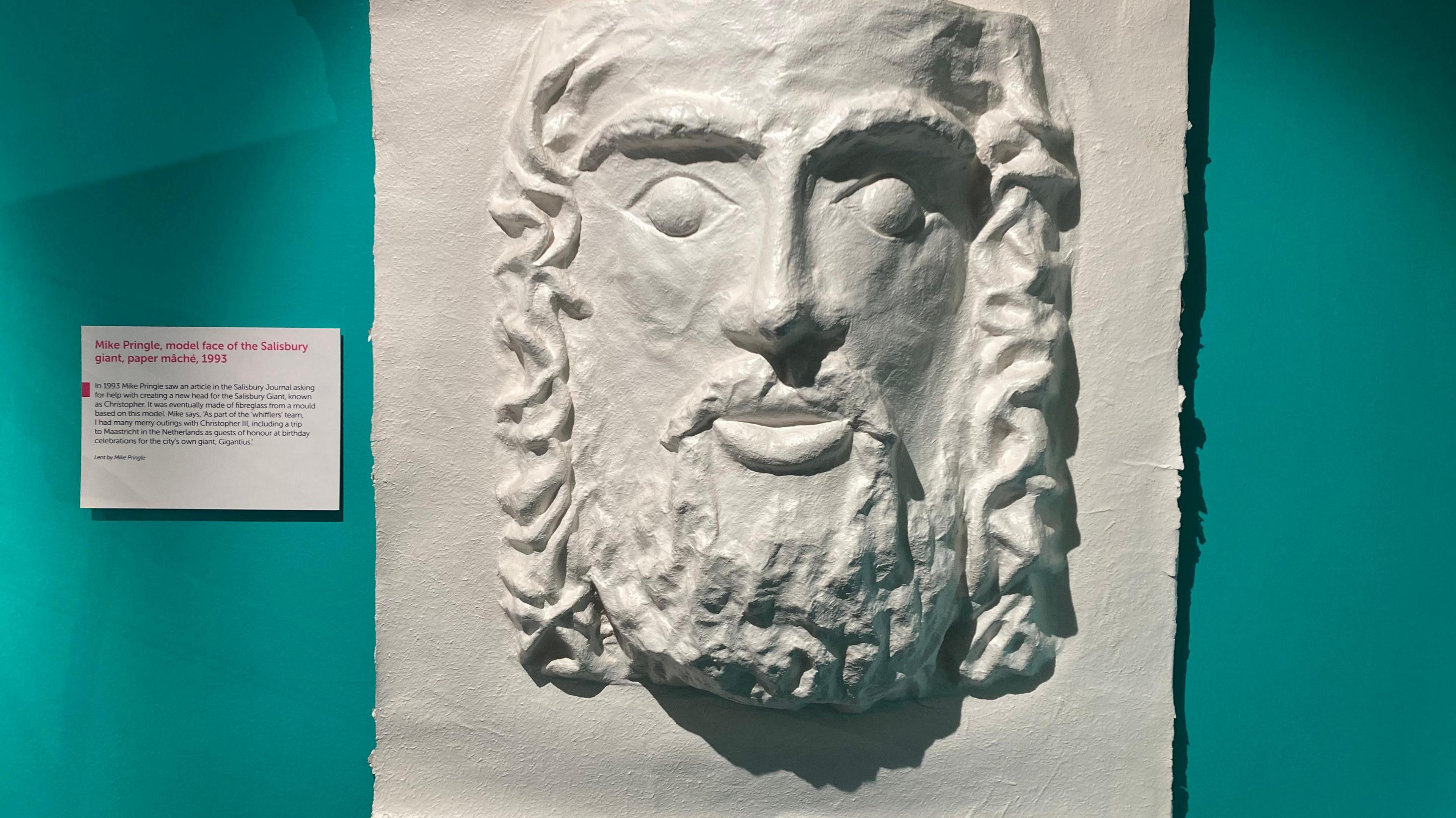 A white plaster-cast style sculpture on the wall of the face of a bearded man. Plaque at the side says it is made by Mike Pringle.