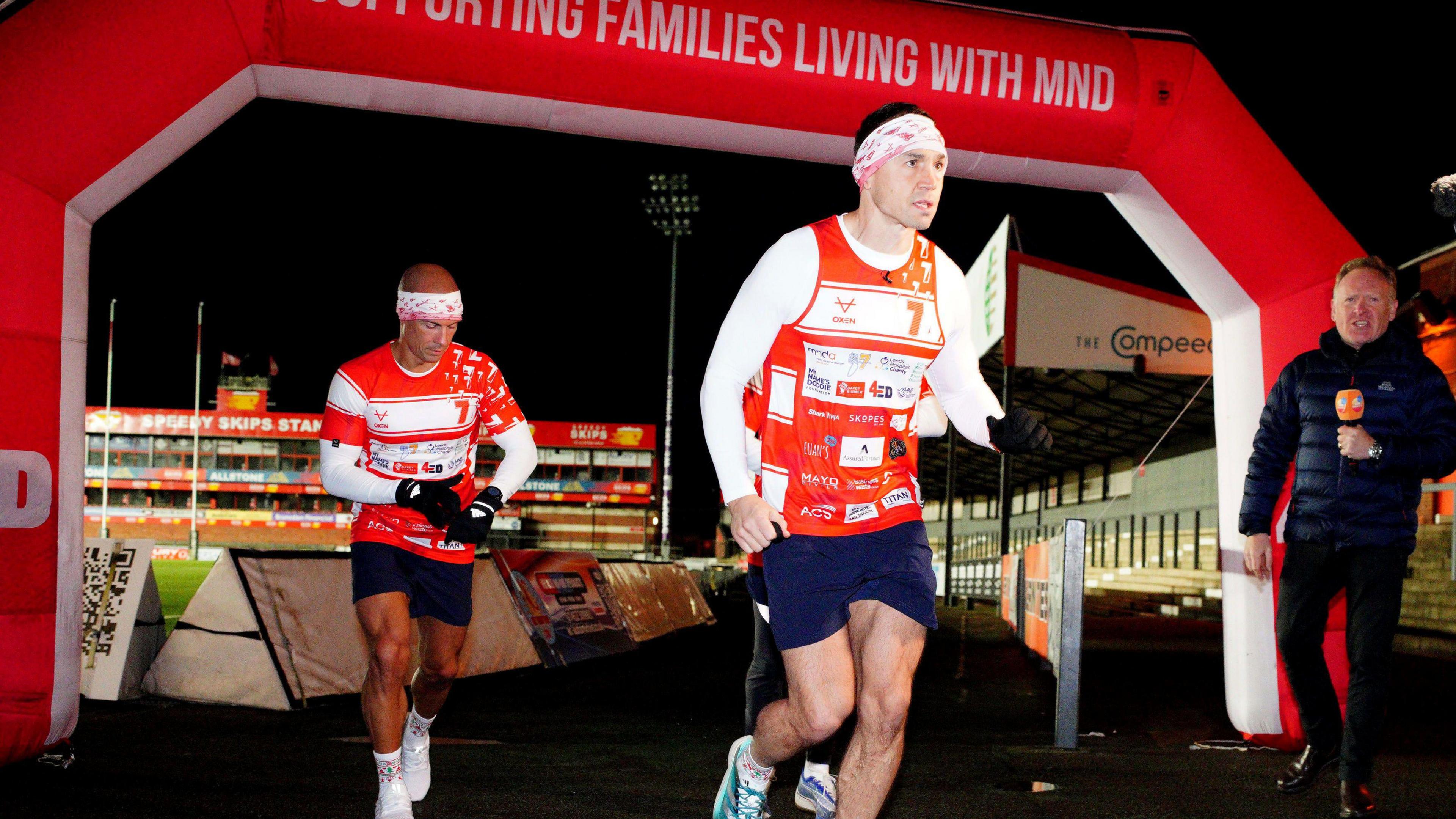 Kevin Sinfield wearing a white top and red vest with the name of charities across it. The picture shows him setting off at the starting line, under an MND arch, with the stadium visible in the background. 