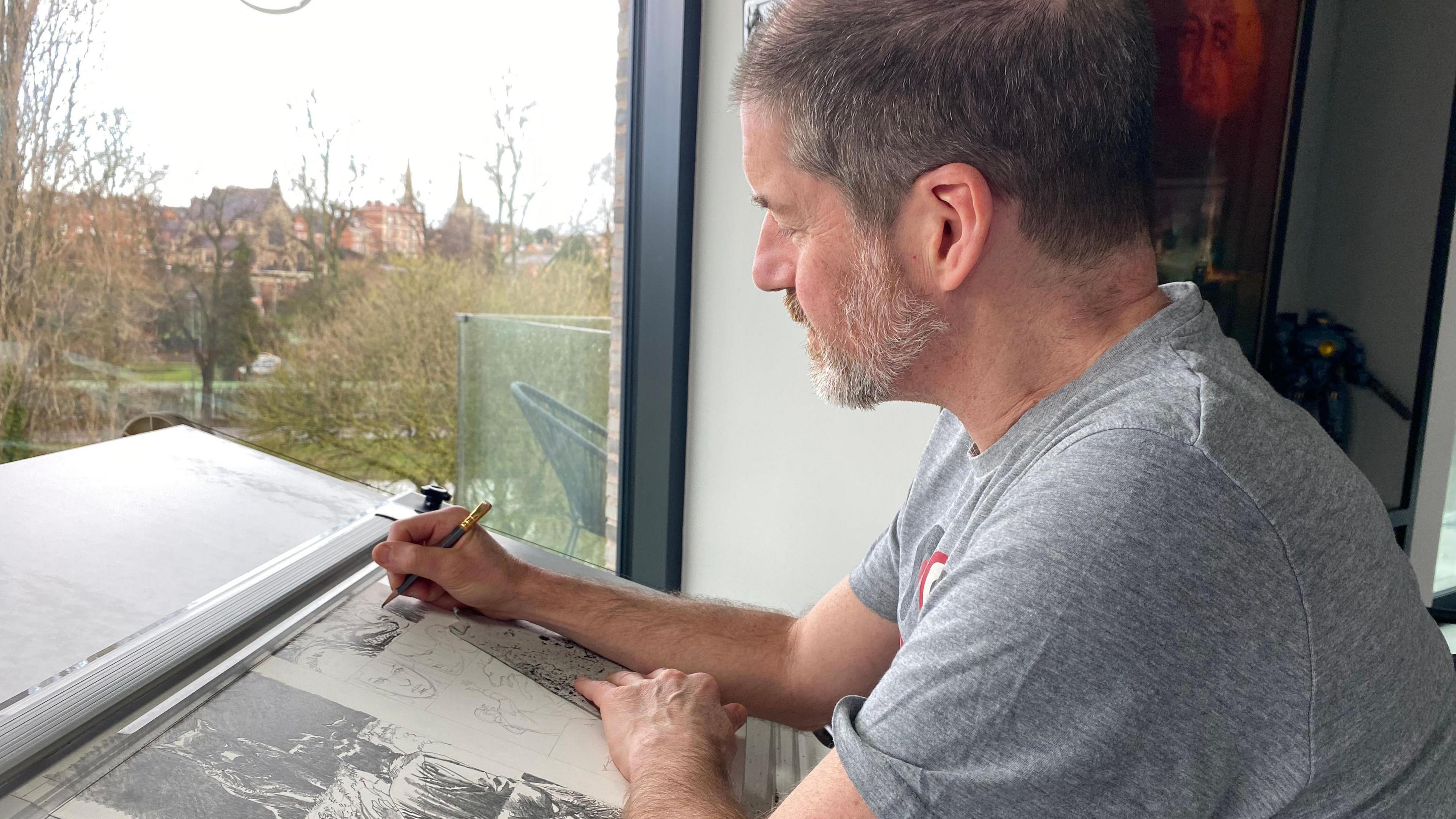 Charlie Adlard sat at his sketching desk, the photo is taken side on so he's pictured in profile. On his desk is a work in progress - one of the images is a wolf. Out of the window you can just about see the River Severn, with trees alongside it. The sky is white and it's recently snowed. 