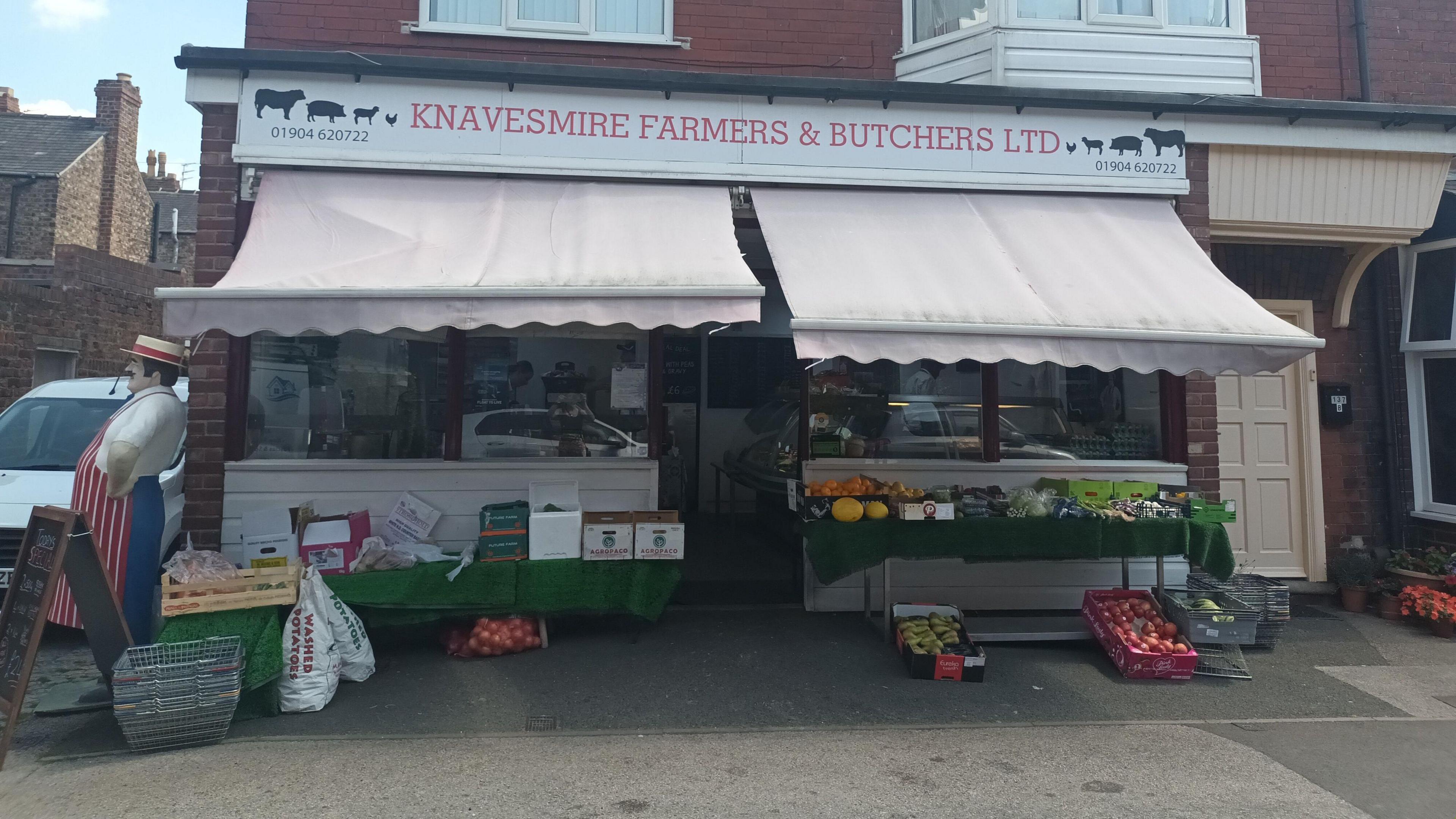The exterior of a traditional butchers shop, with a white fabric awning and an outdoor display of fruit and vegetables. 