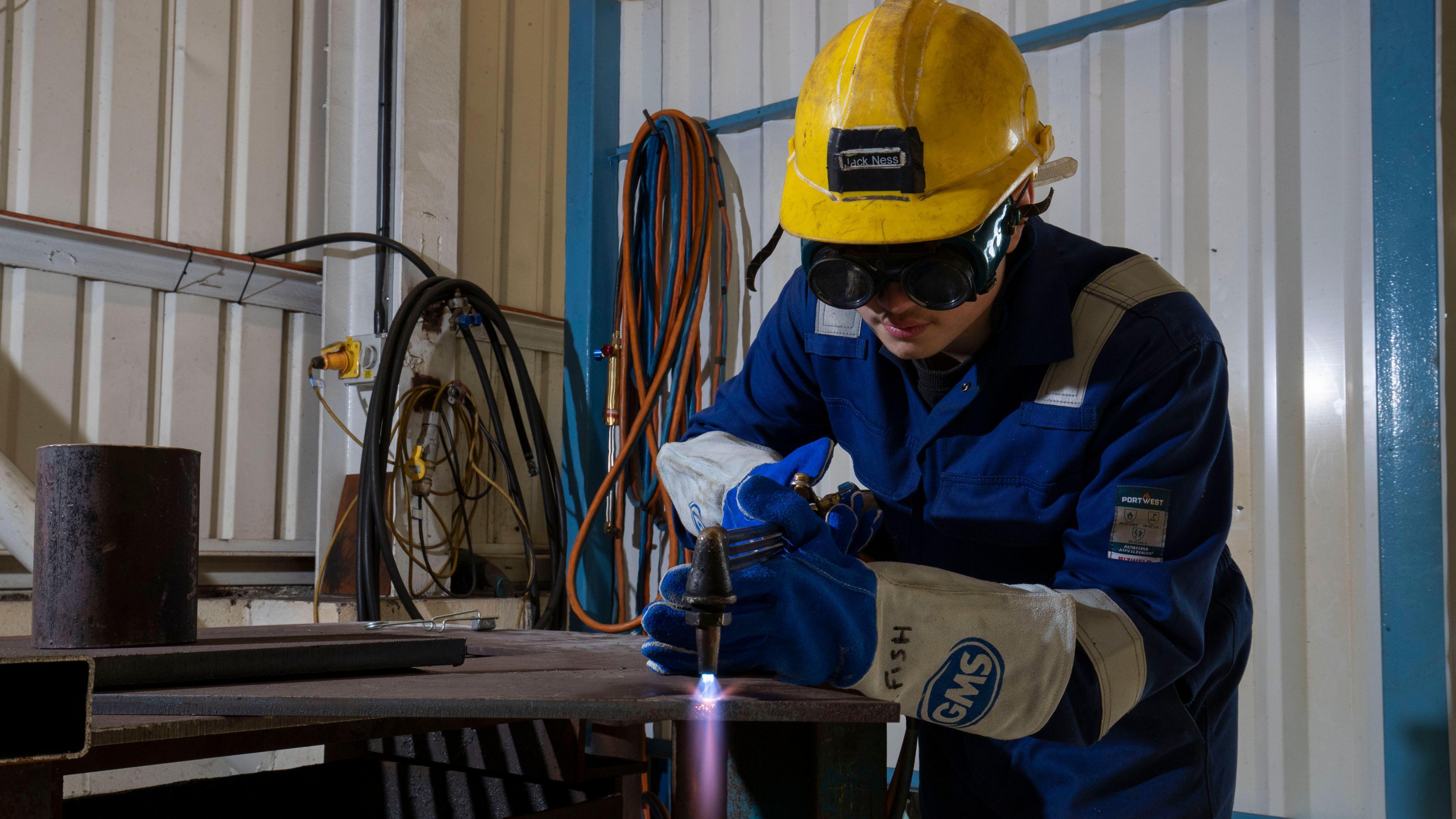 A man in working gear with protective glasses and a helmet is using what appears to be a blow torch or a welder 