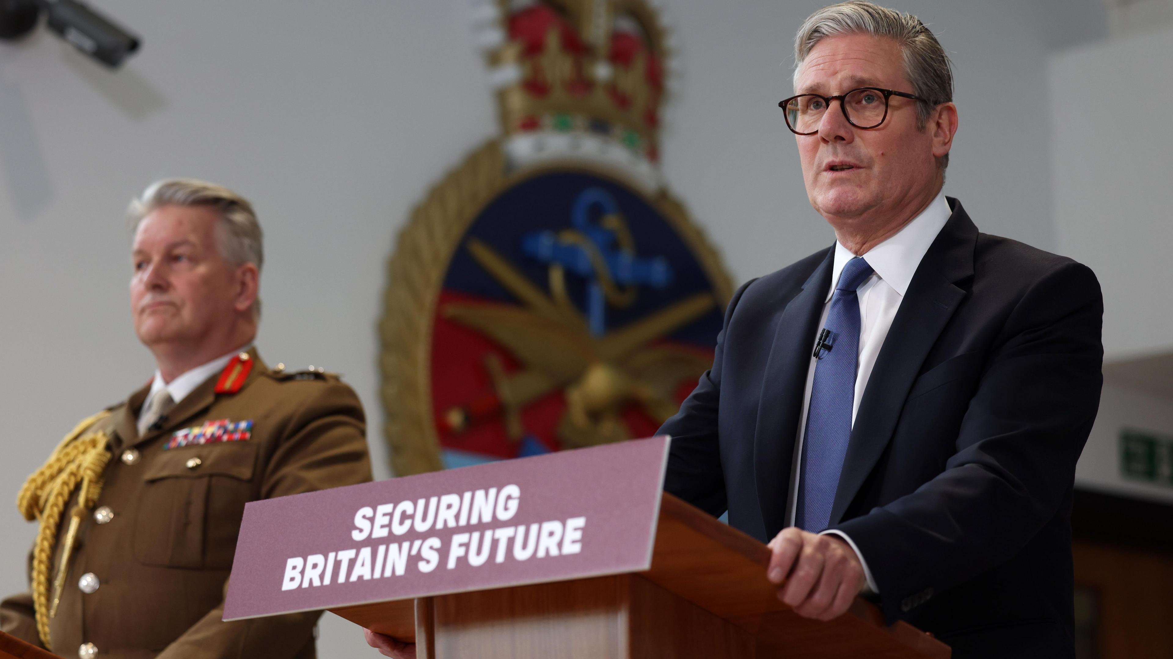 UK Prime Minister Keir Starmer in a suit, accompanied by General Sir Jim Hockenhull in military uniform standing at podiums with a sign that reads 'Securing Britain's Future'