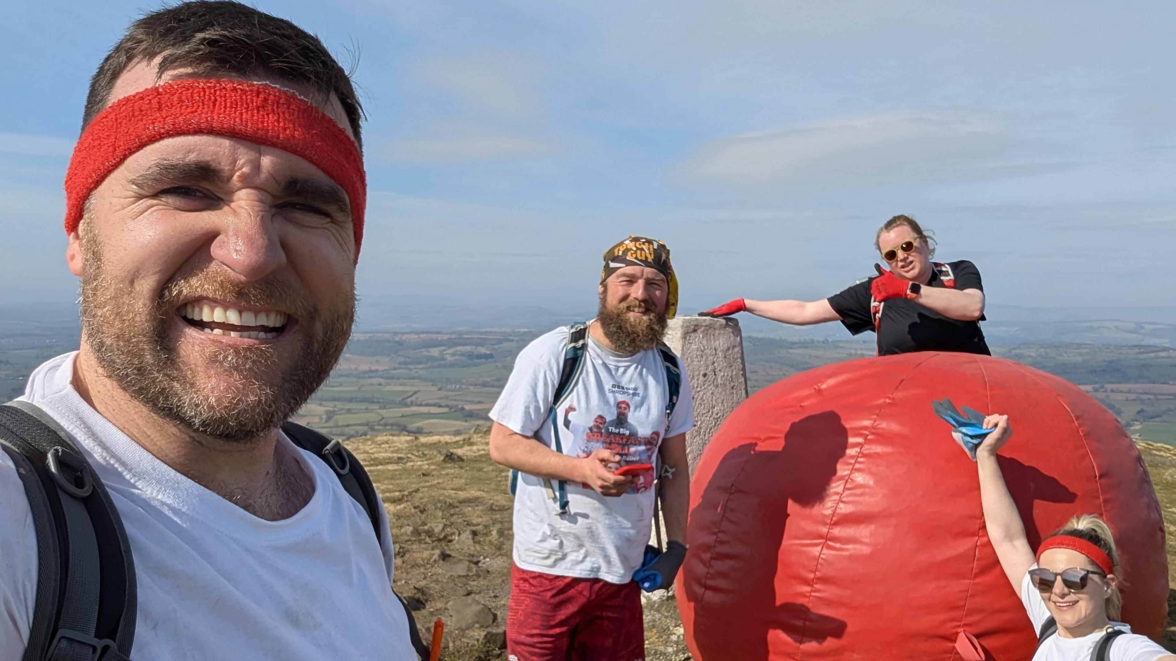 Tim Barnes has short brown hair and is wearing a red headband. He is taking a selfie on top of a hill. In the background are Adam Green, who has a large beard and is wearing red and white, Carlie Swain, who is standing behind a large red ball with her thumb up, and Michaela Wylde, who is crouched beside the ball with her arm in the air.