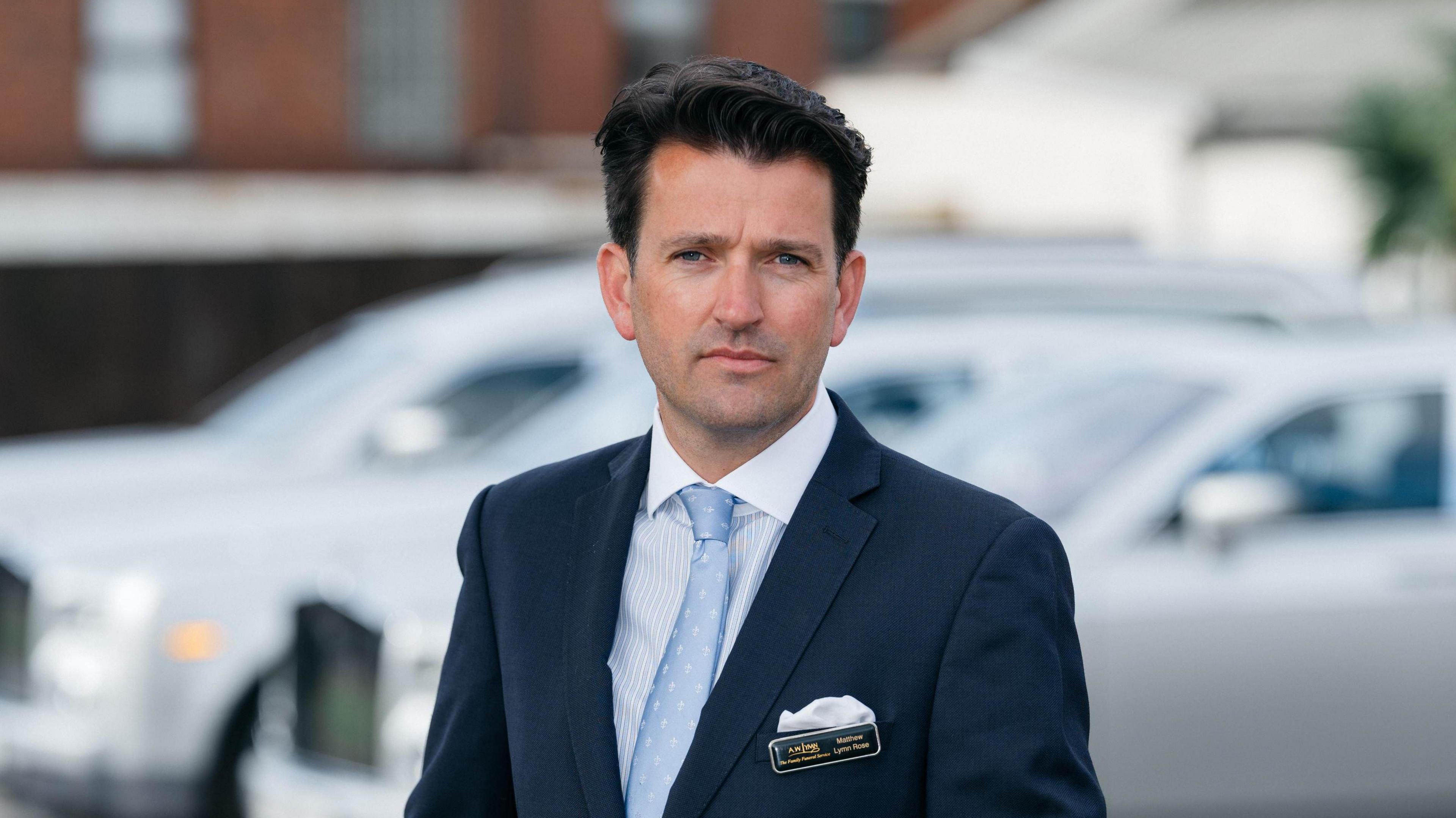 Matthew Lymn Rose with dark hair, wearing a suit and light blue tie