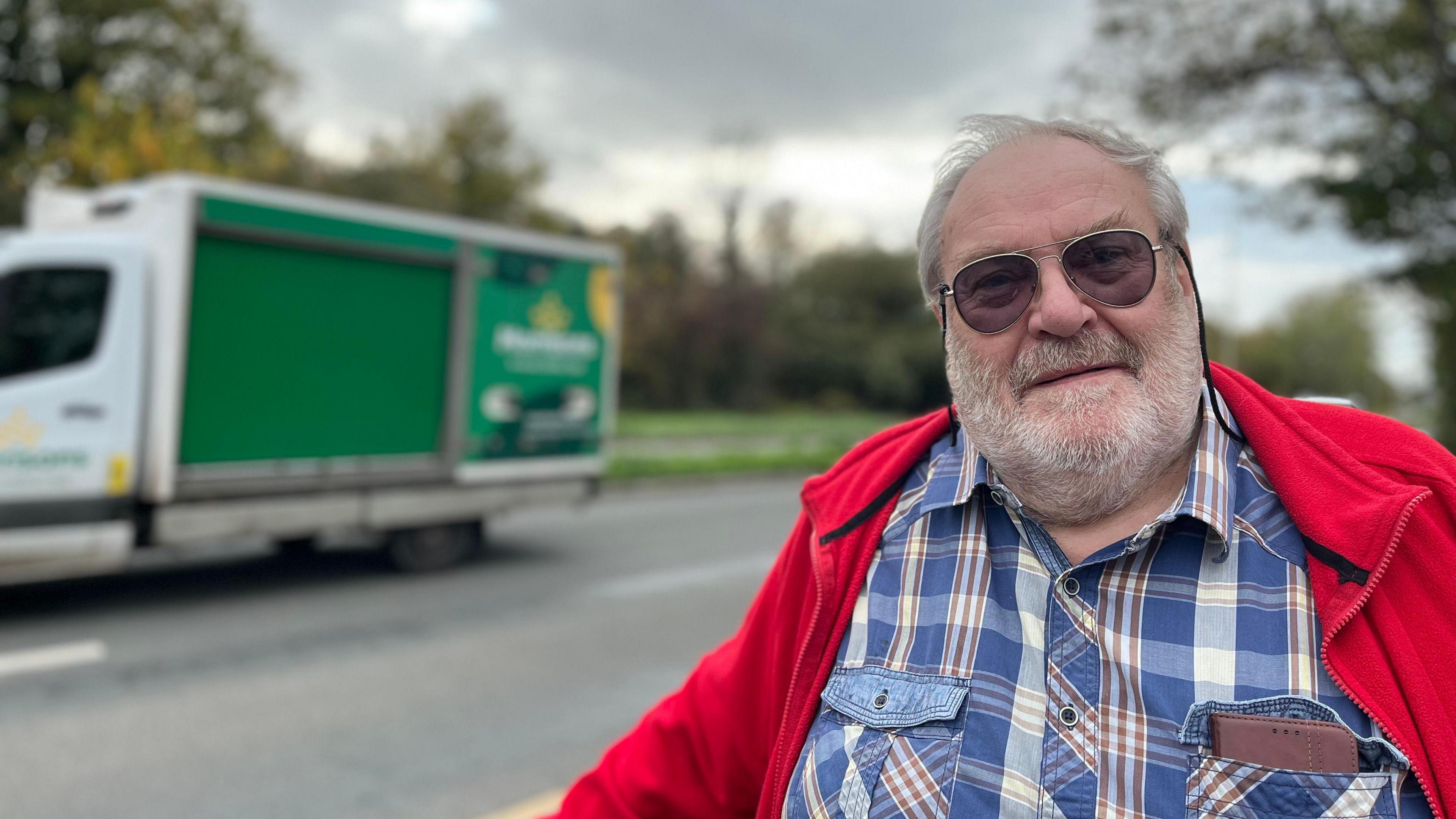 Chris Greenwood looks at the camera - he's wearing sunglasses, a red fleece and blue check shirt. He has a white beard. Blurred behind him is a duel carriageway, with a large van going past