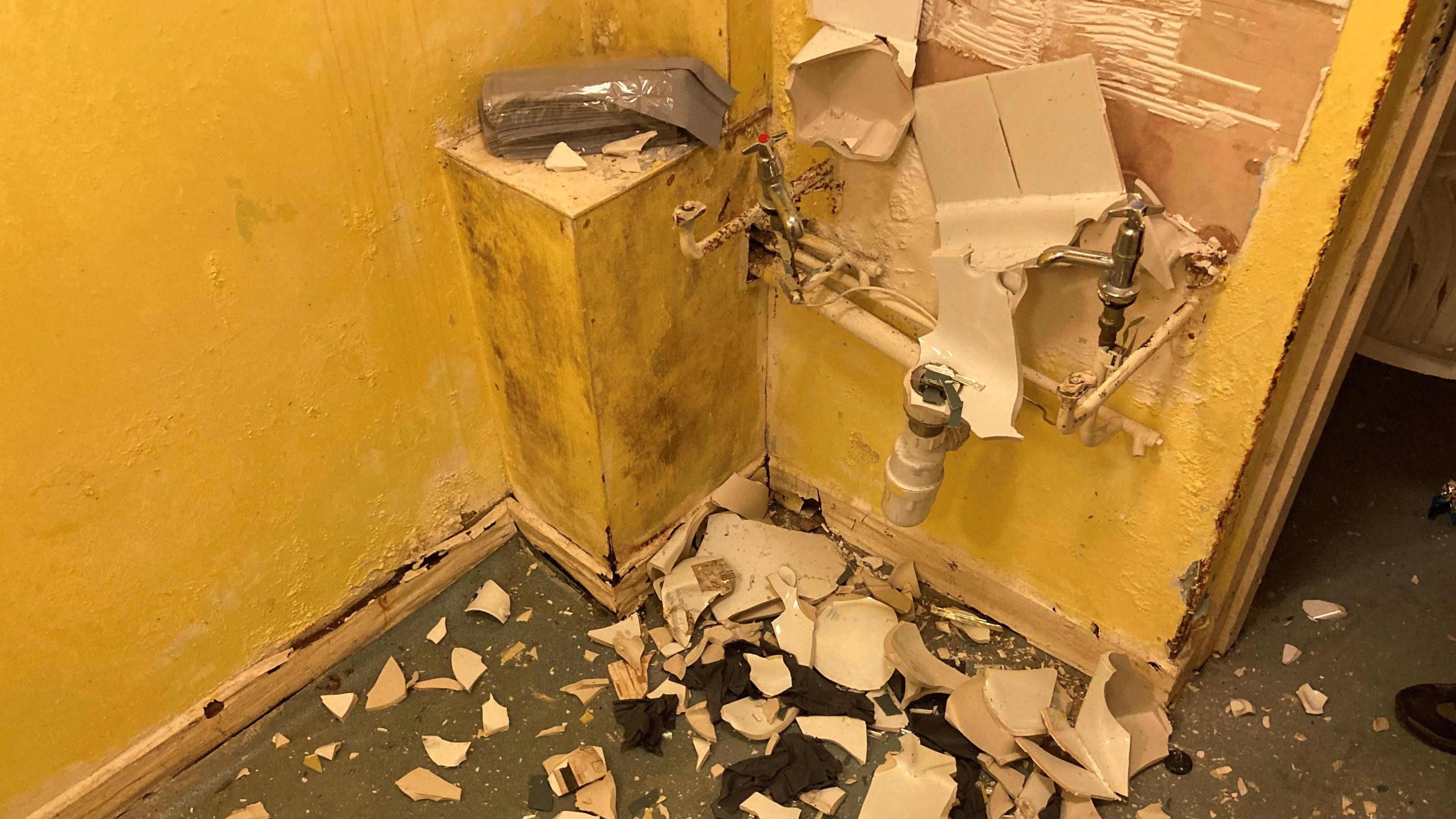 A wall showing what used to be a sink, broken into white pieces on the ground