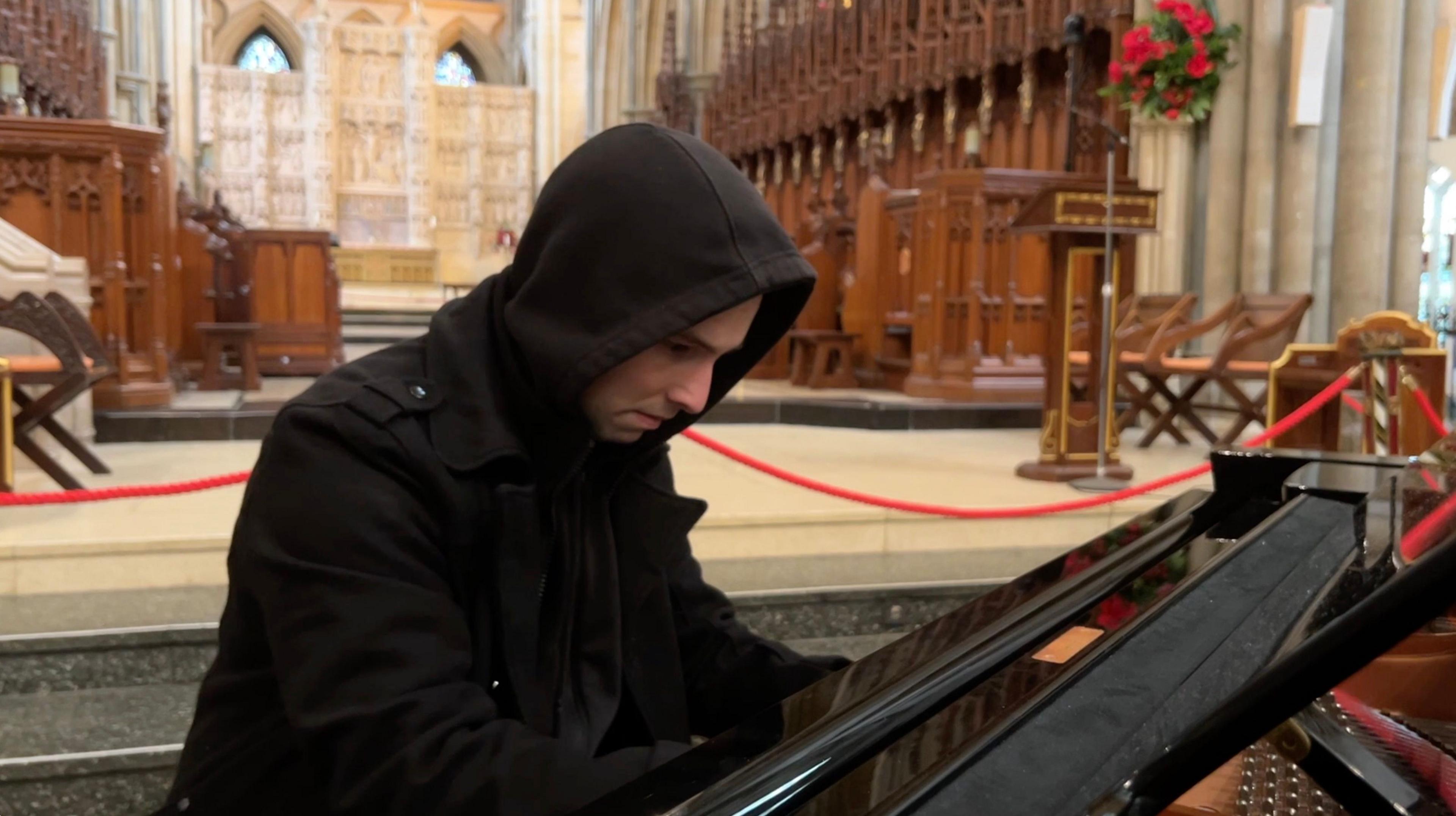 A man in a black hooded coat plays the piano in a church.