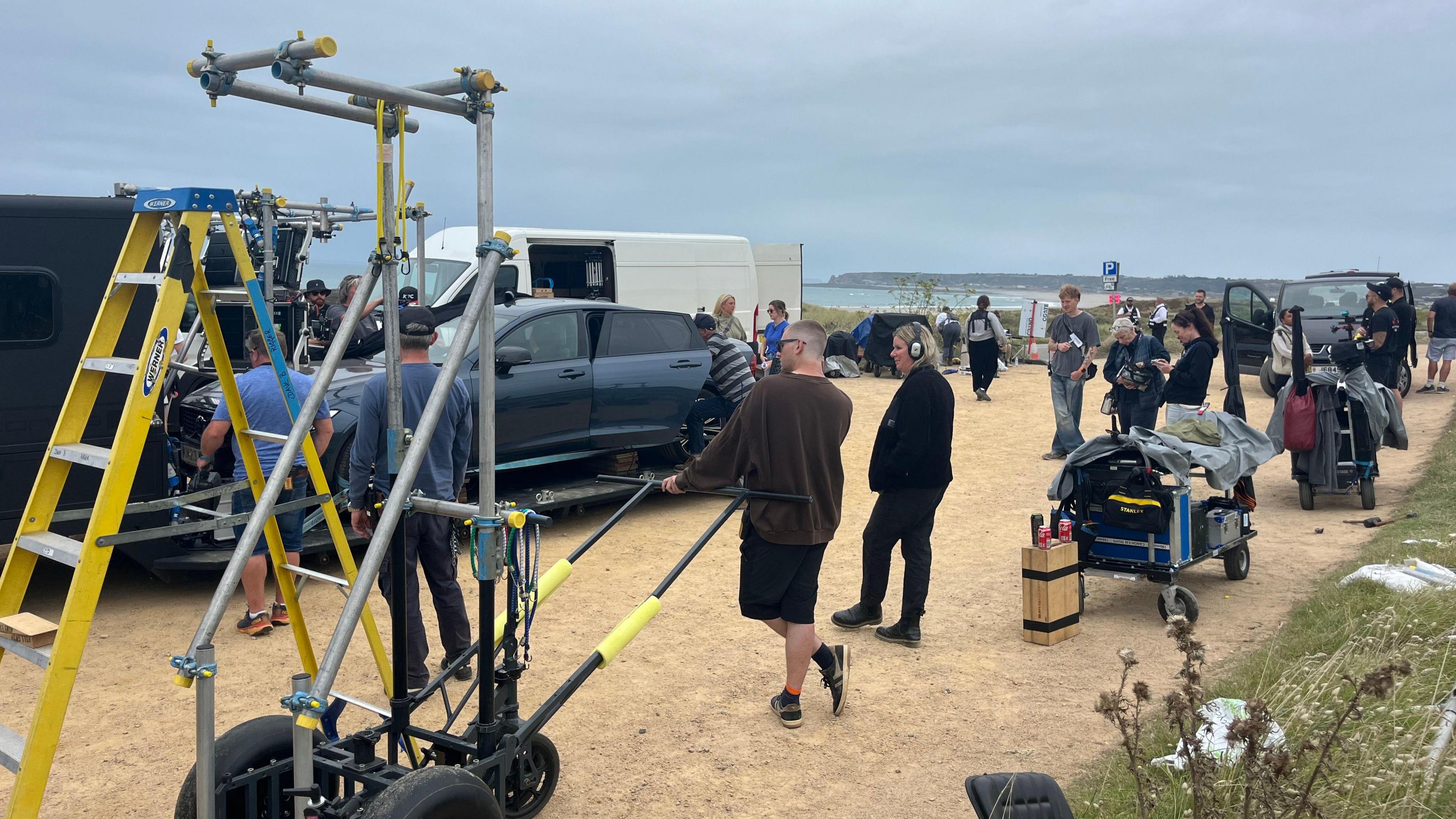 A film crew is operating a car rig in a sandy car park next to a main road along St Ouen's Bay. 