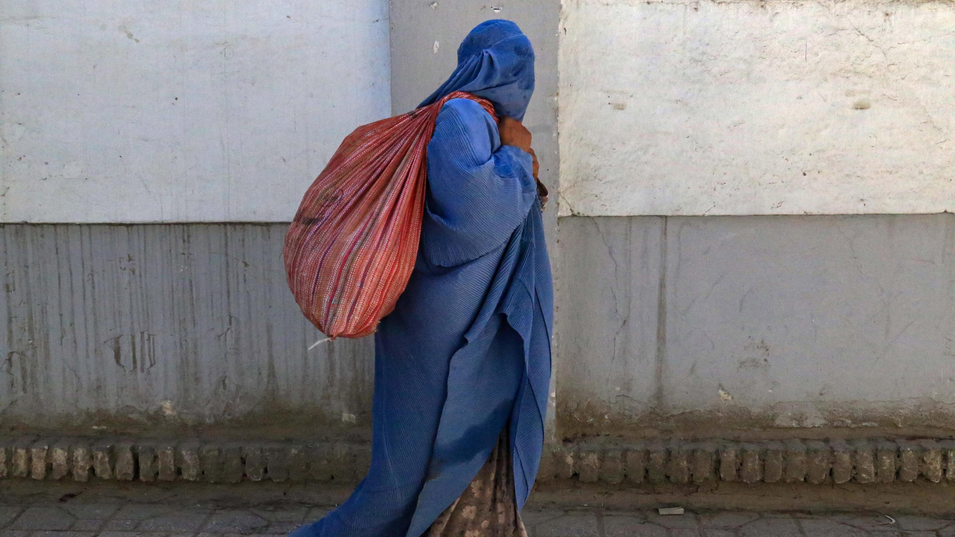 A woman wearing a blue burqa walks down a street in Kabul with a red sack over her shoulder. You cannot see any of her face