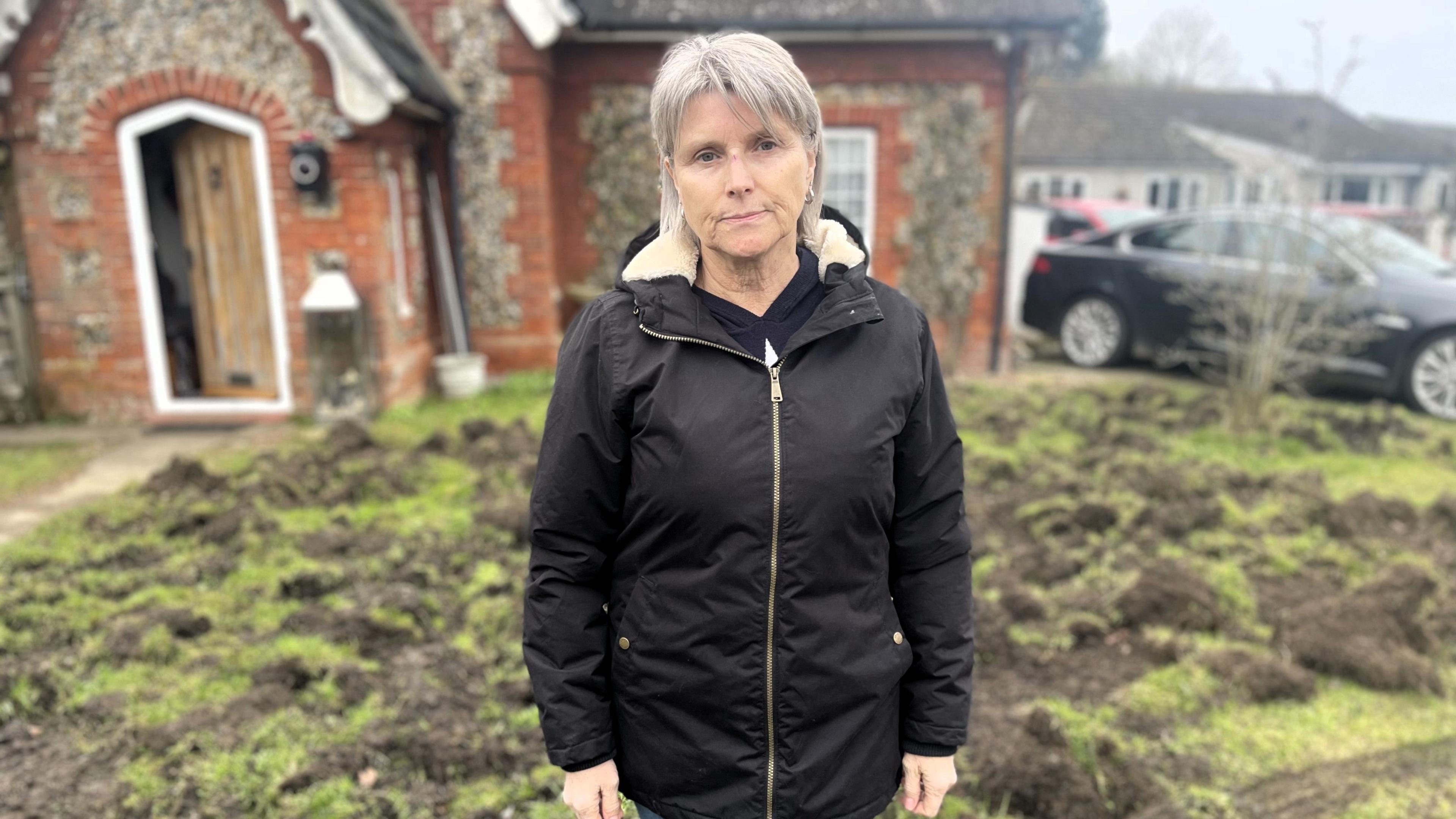 Caroline Harris, with grey hair and a black jacket, standing by her heavily damaged lawn.