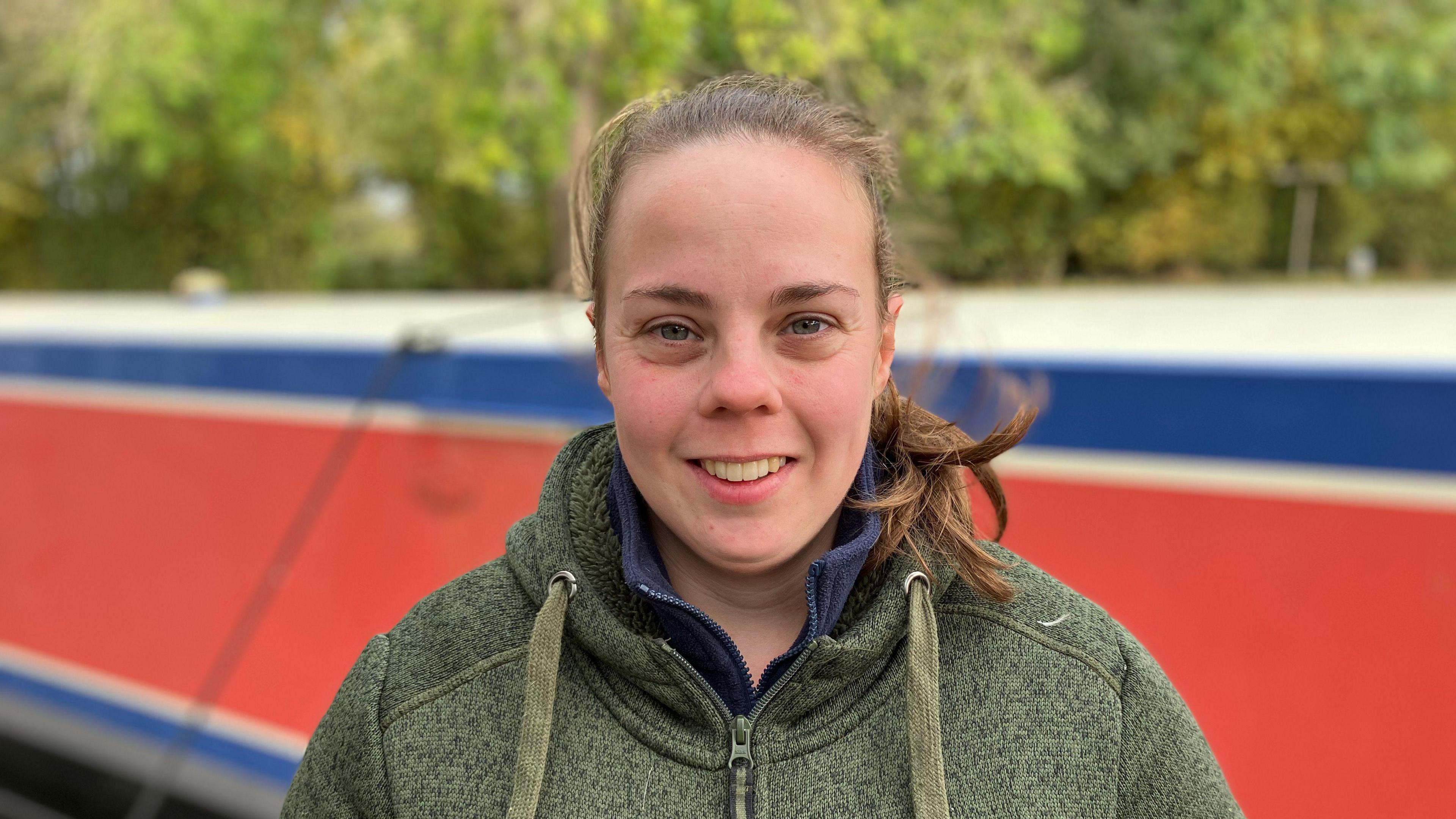 Rebecca Howard is standing facing the camera smiling in front of her red boat and she is wearing a dark green fleece.