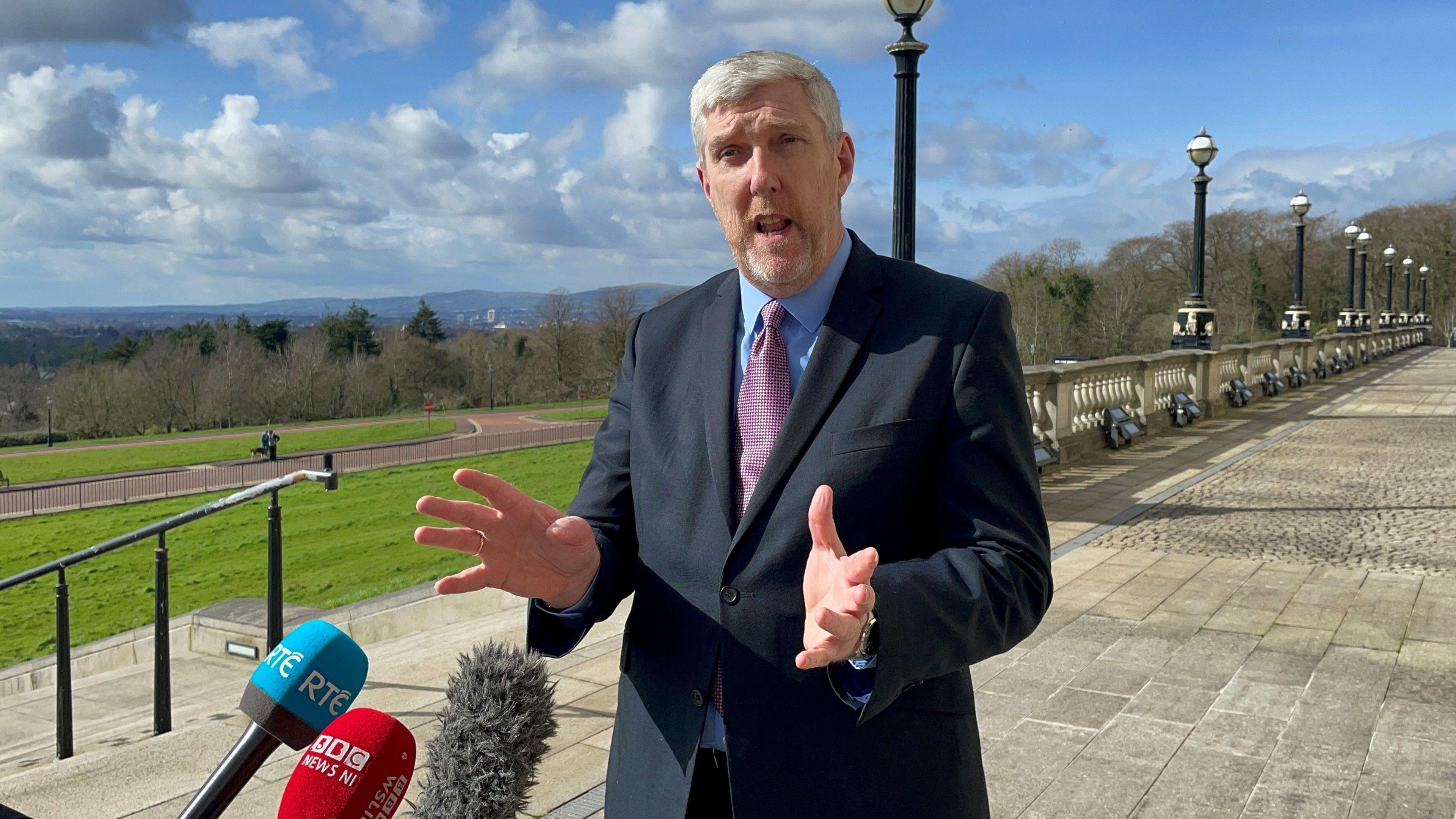 Infrastructure Minister John O'Dowd standing behind media microphones. He is standing outside Parliament Buildings in Stormont, with the green area in the background