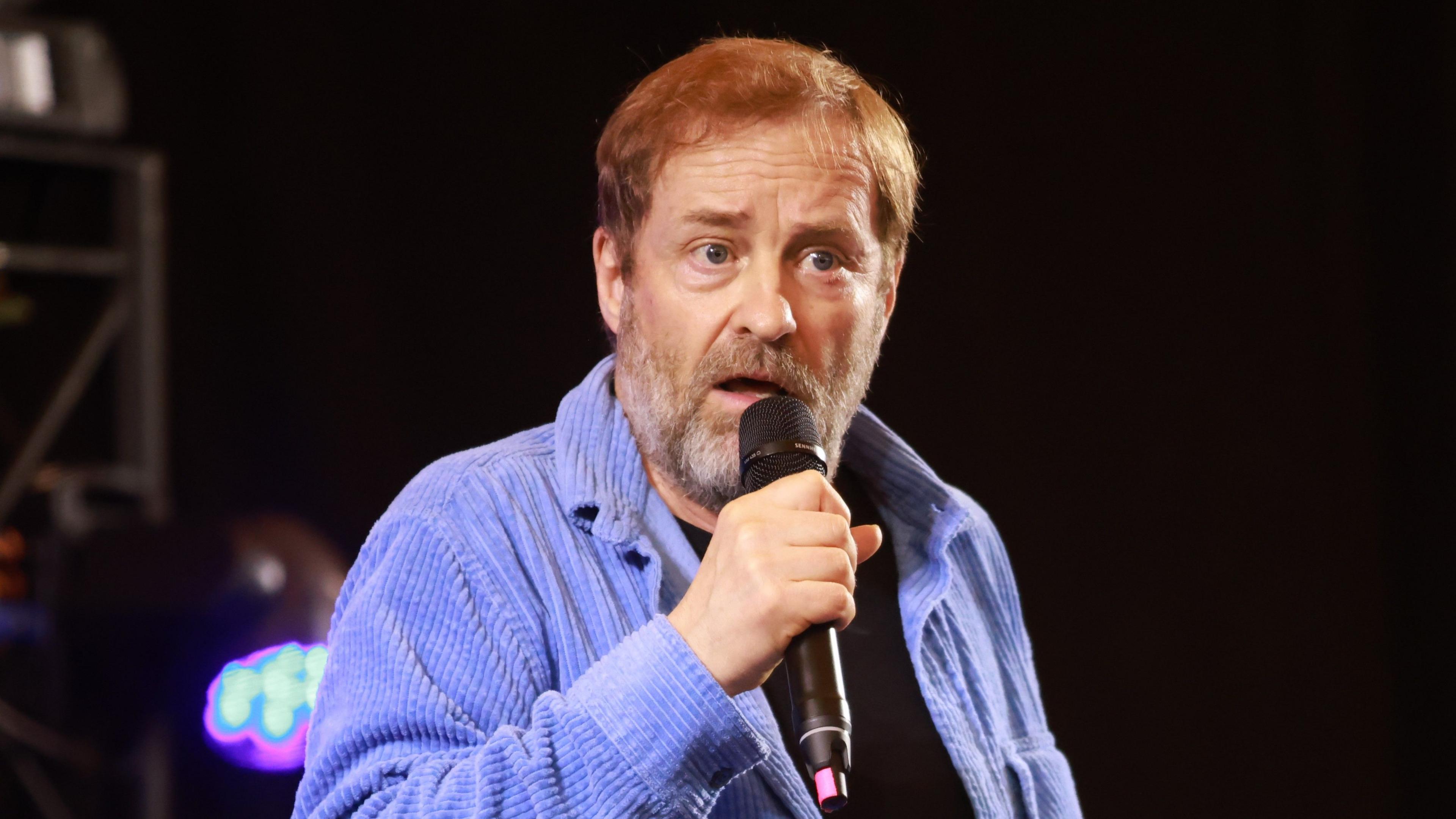 The comedian Ardal O'Hanlon holding a microphone while performing on stage. He has a grey, short beard, short brown hair and is wearing a black t-shirt and a purple corduroy shirt.