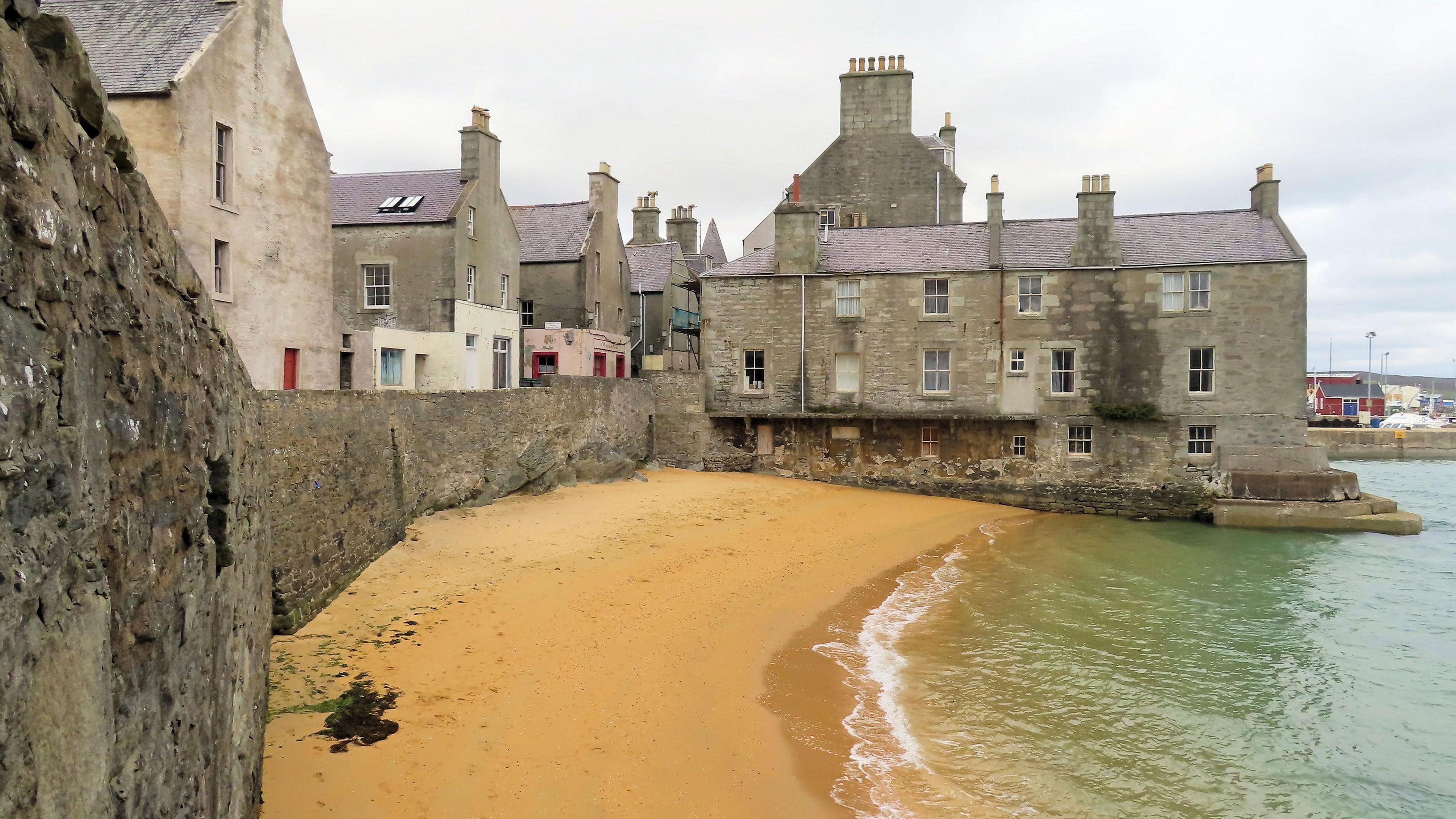 Orange sand against a green sea with a grey house jutting out over the sea. There is a long sea wall and houses on the left of the picture.