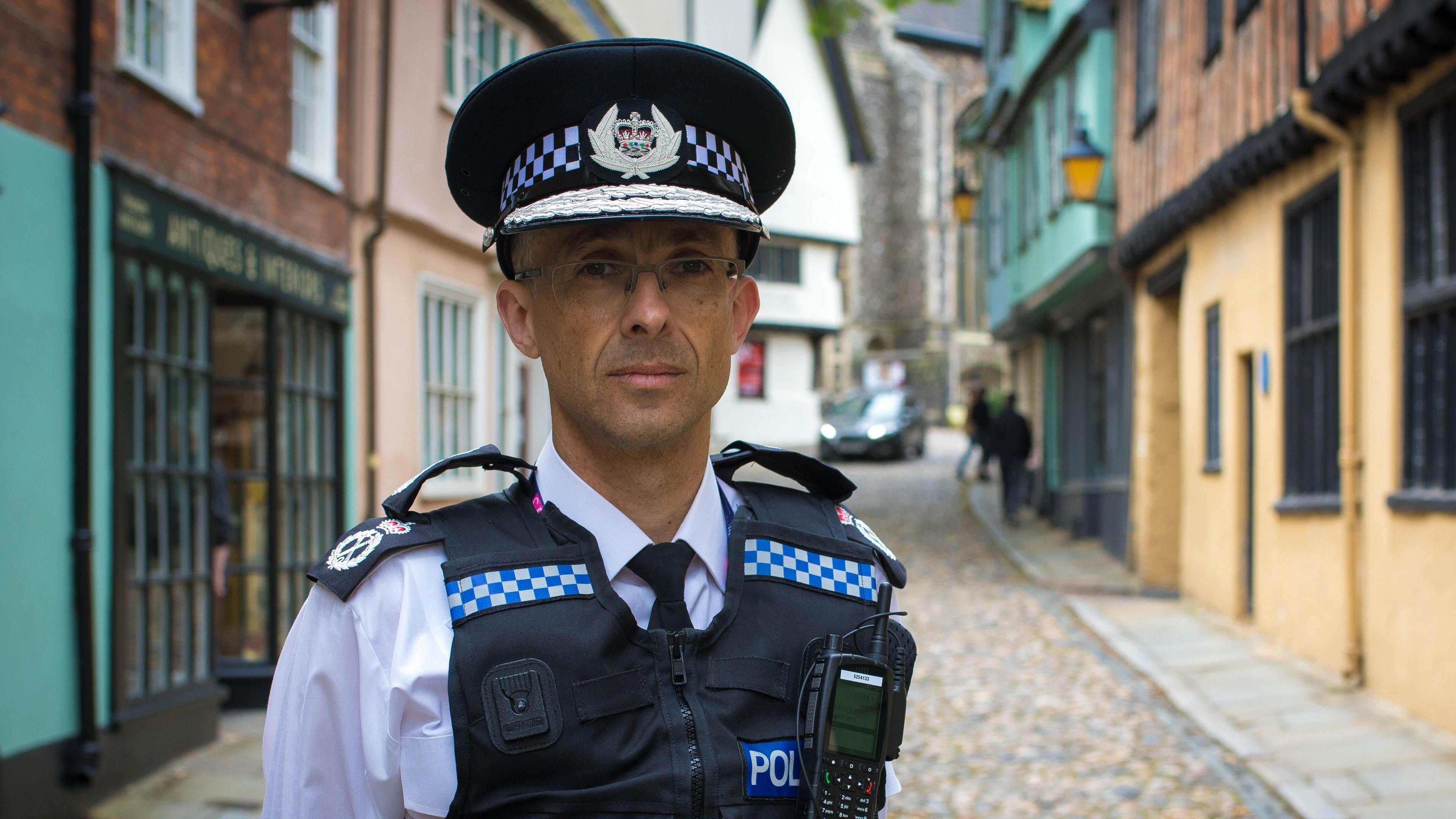 Chief Constable Paul Sanford standing in uniform on a cobbled street in Norwich. He is wearing glasses and a peaked hat