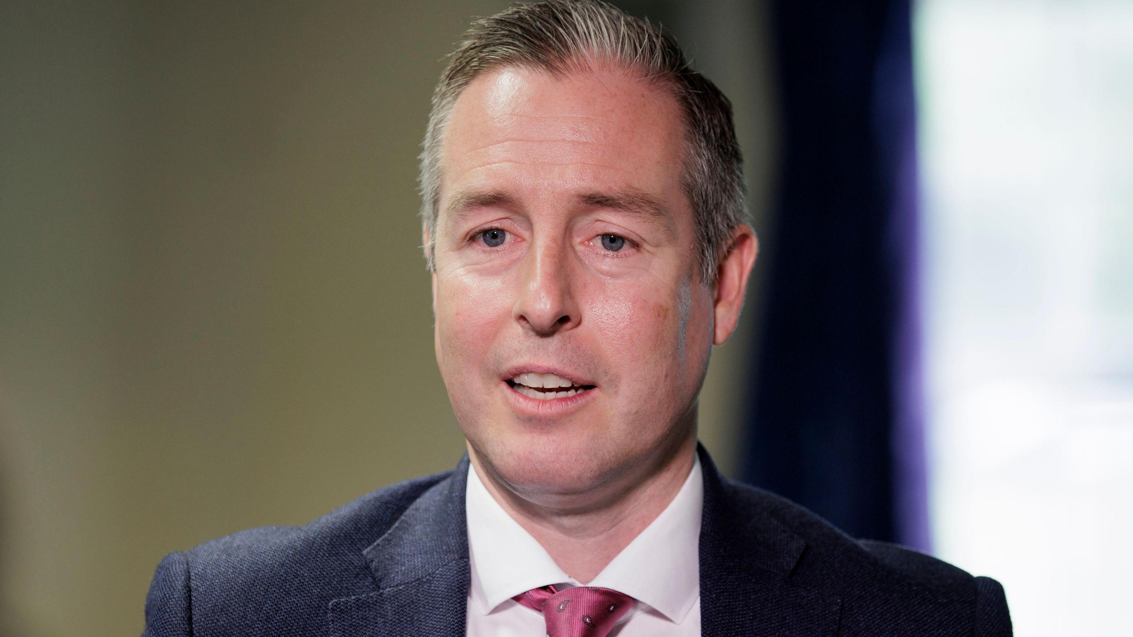 Paul Givan, a man with short brown hair, a dark suit jacket, white shirt and red/pink tie