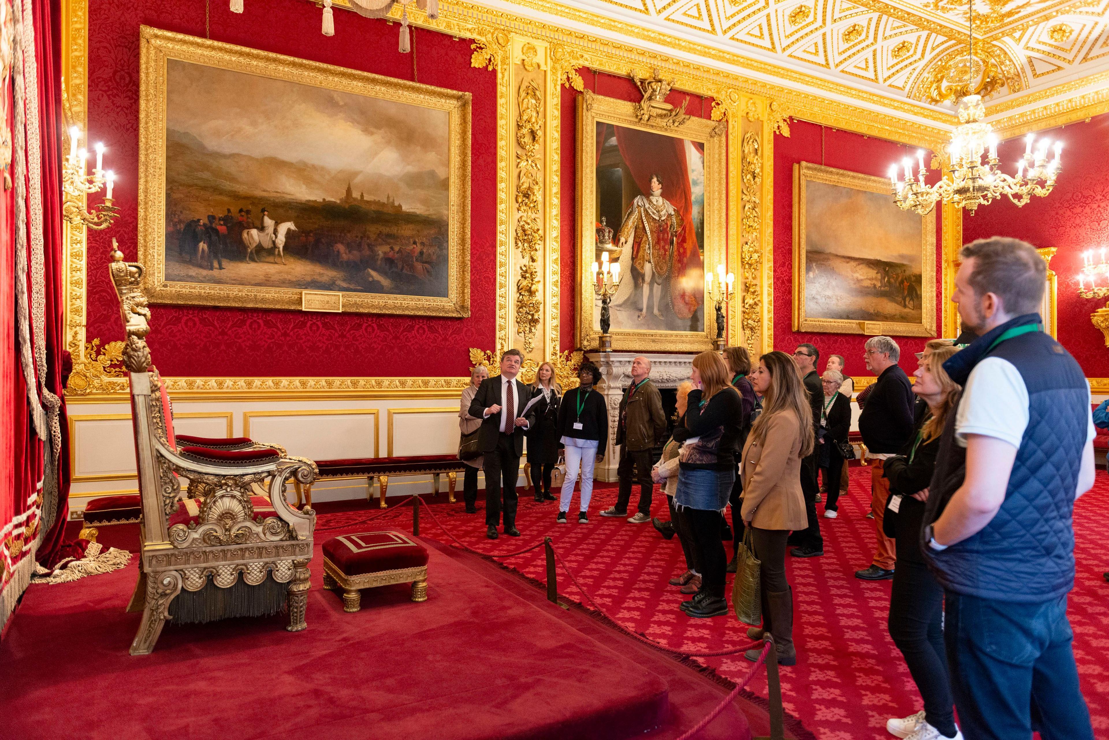 A group of tourists and their guide stand in front of a throne in the throne room, which is richly decorated in red and gold. There are monumental paintings on the wall. 