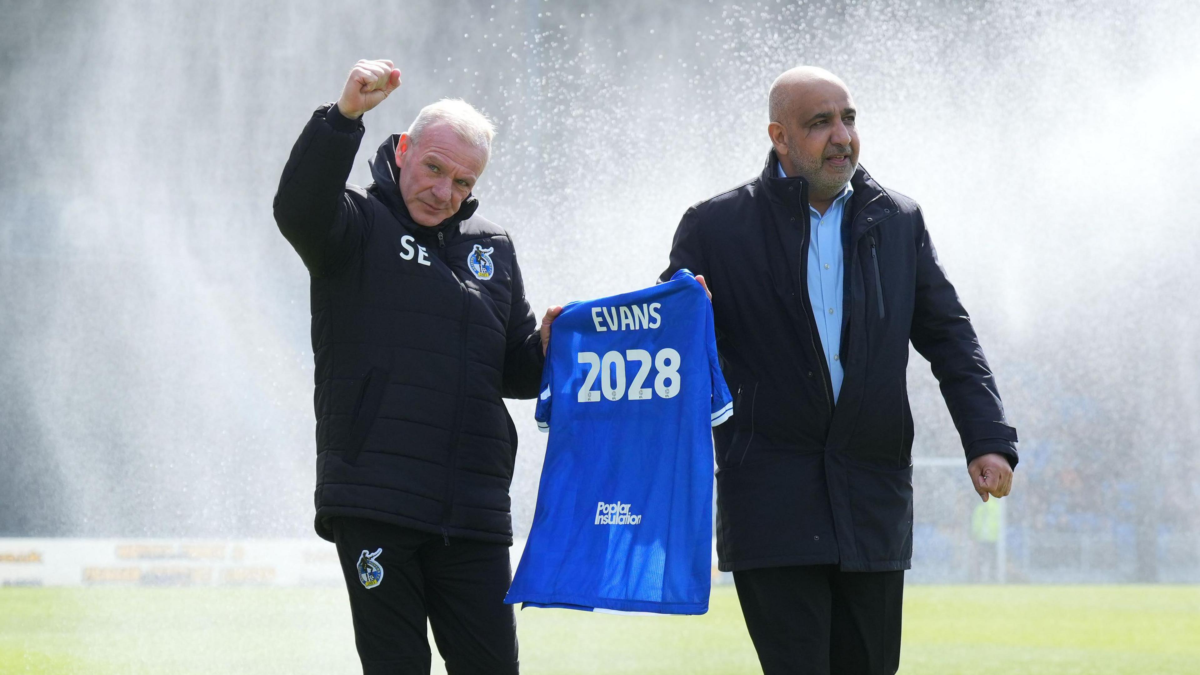 Steve Evans (left) walks on the pitch with his right arm in the air and fist clenched and his left hand holding a Bristol Rovers shirt with 'Evans 2028' written on the back. Hussain AlSaeed (right) walks alongside Evans holding the shirt with his right hand