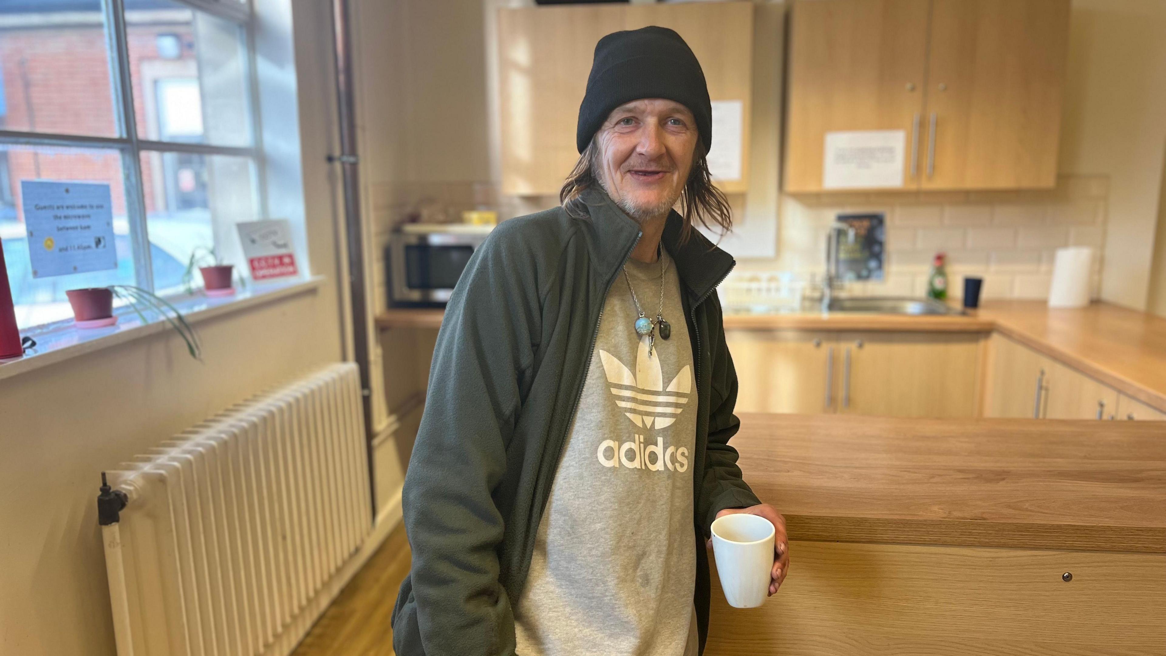 Man wit long grey hair leans on kitchen service holding white mug, wearing black beanie, grey adidas t shirt and fleece 