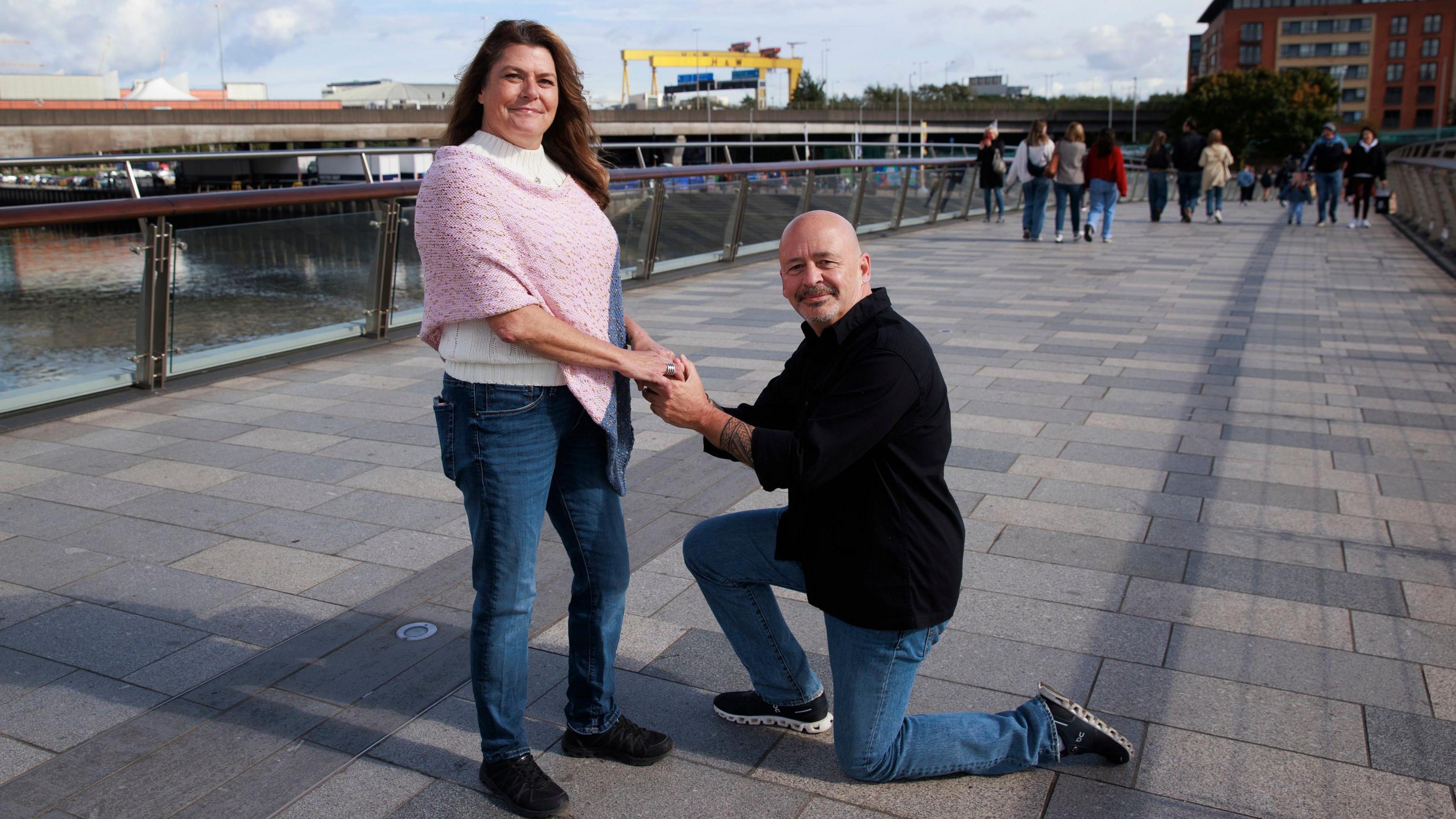 Angela Harsanyi and Gian Perroni on the Lagan Weir. Gian is down on one knee. Angela has long brown hair, wearing a white top, pink knit poncho and jeans. Gian is bald and wearing a black shirt and jeans.