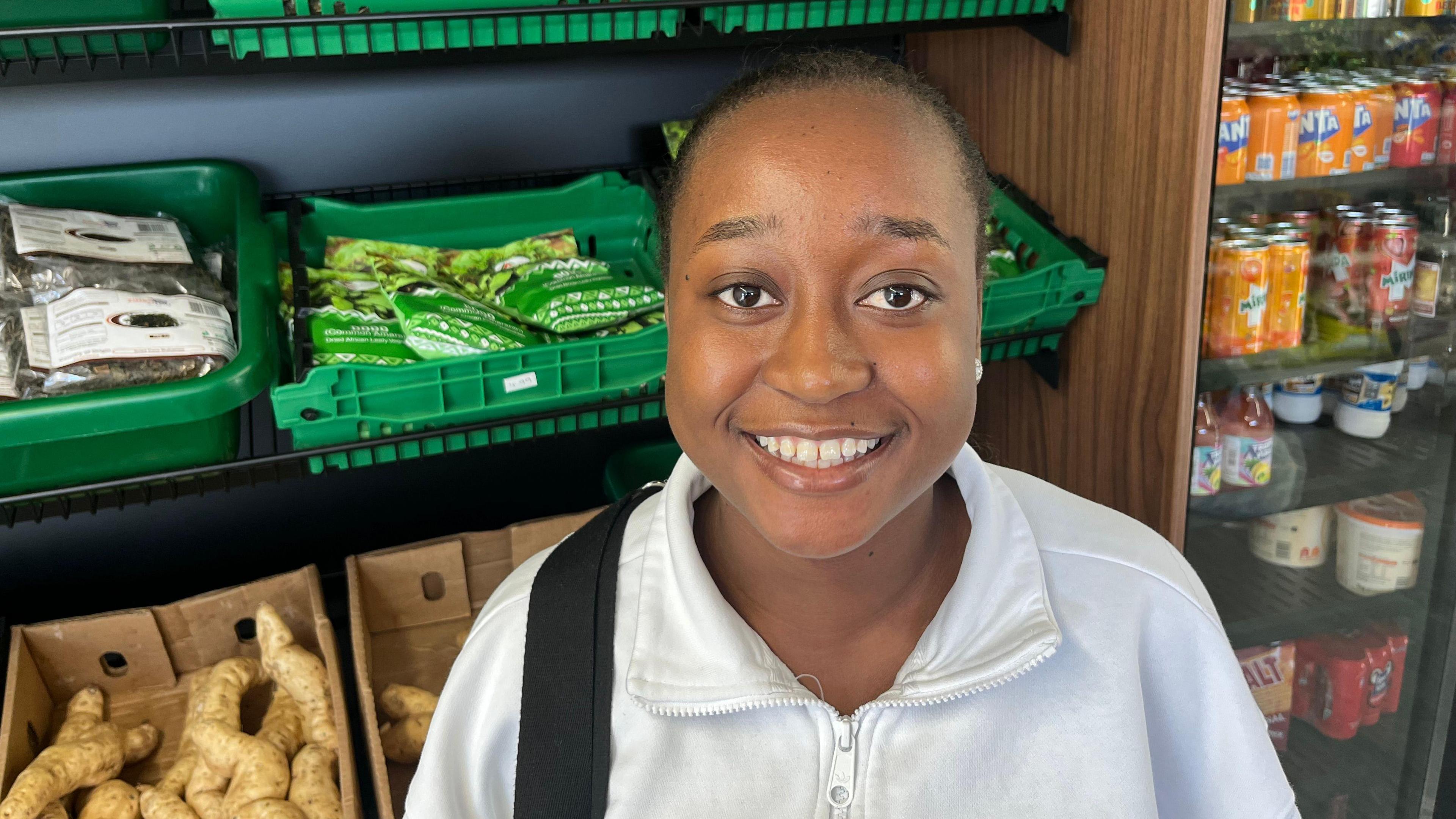 Perpetual Gumbo is standing inside a grocery shop on the farm. She is wearing a white zip-up jacket and has a black strap across her shoulder, possibly from a bag. She is smiling warmly at the camera. Behind her, shelves are filled with green trays holding packaged food items, including dried goods and fresh produce. To the right, there’s a refrigerated section stocked with drinks, including brightly coloured cans and bottles from Zimbabwe.