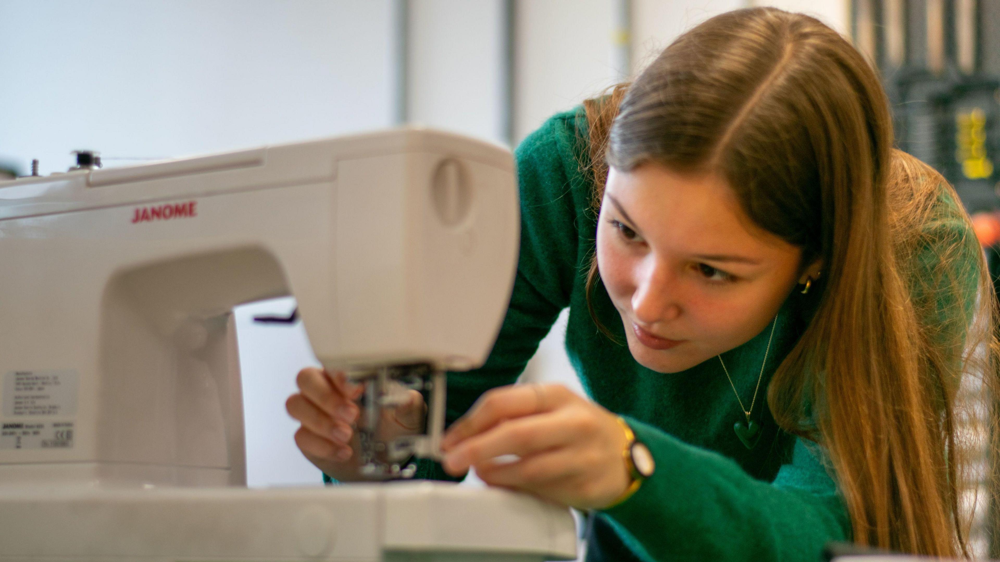 Pascha is pictures bending down to a white sewing machine. She is wearing a forest green jumper. 