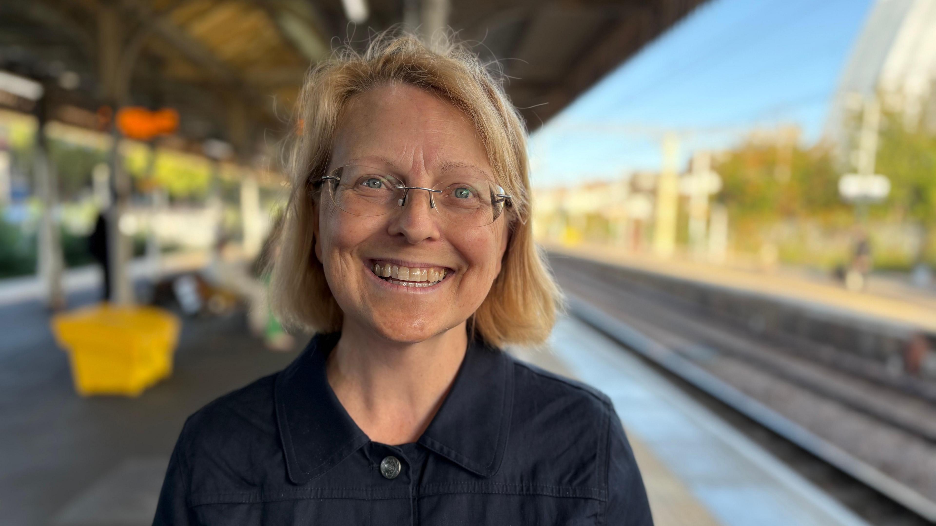 A woman with blonde hair and glasses smiles at the camera. She is wearing a dark blue top with a button collar. A railway platform is in the background.