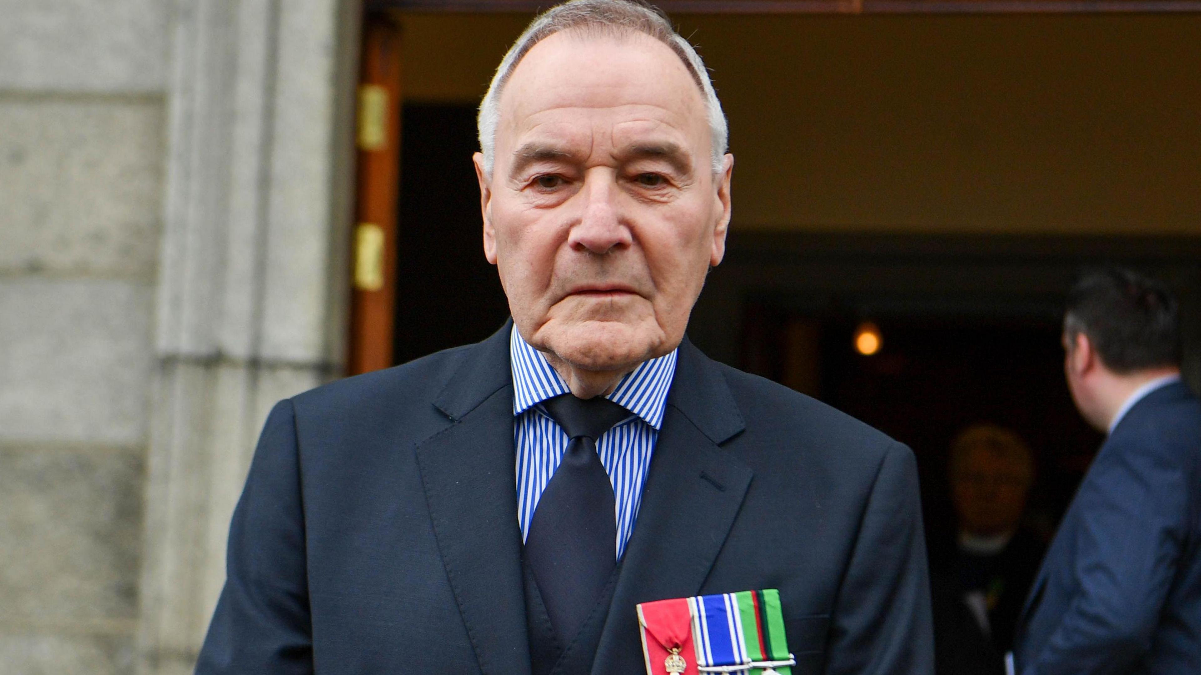 Bill has a navy blue suit on with a blue and white striped shirt and navy tie on. He has grey hair and is wearing three medals -  they are red, blue and white and green and white. 