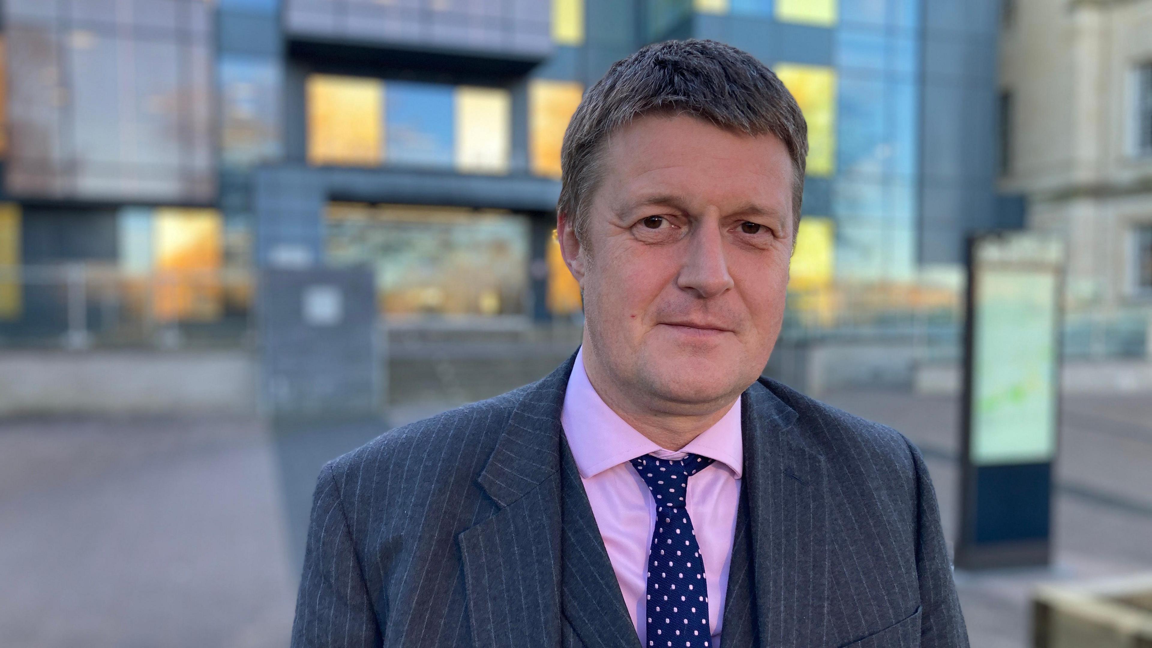 Richard Clewer, pictured outside County Hall. He wears a three-piece grey pinstripe suit with a pink shirt and a blue and pink tie, and has short light brown hair. 