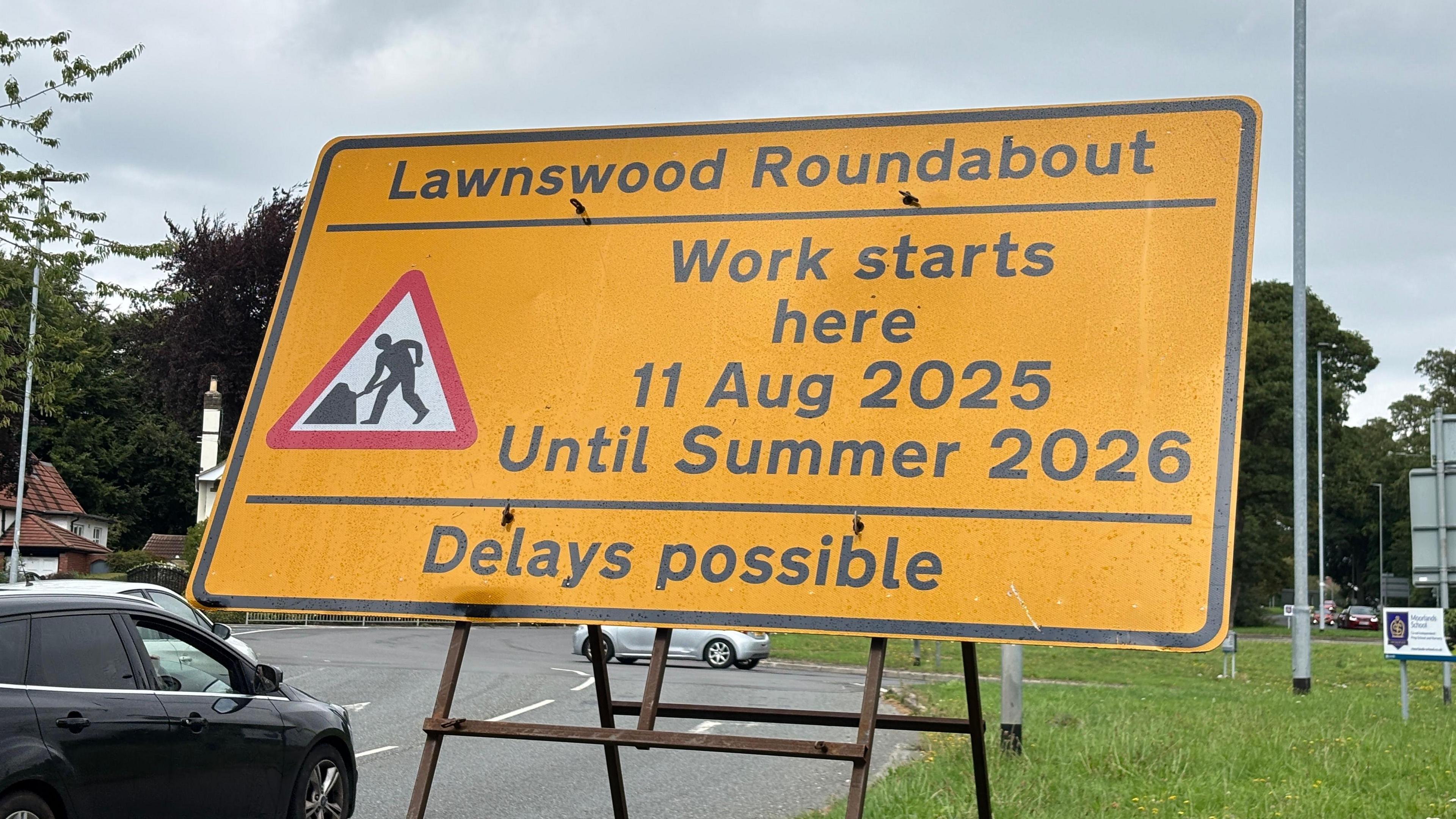 A yellow roadworks sign in front of the roundabout. It says "work starts here" on 11 August 2025, and will last until summer 2026. It says delays are possible.