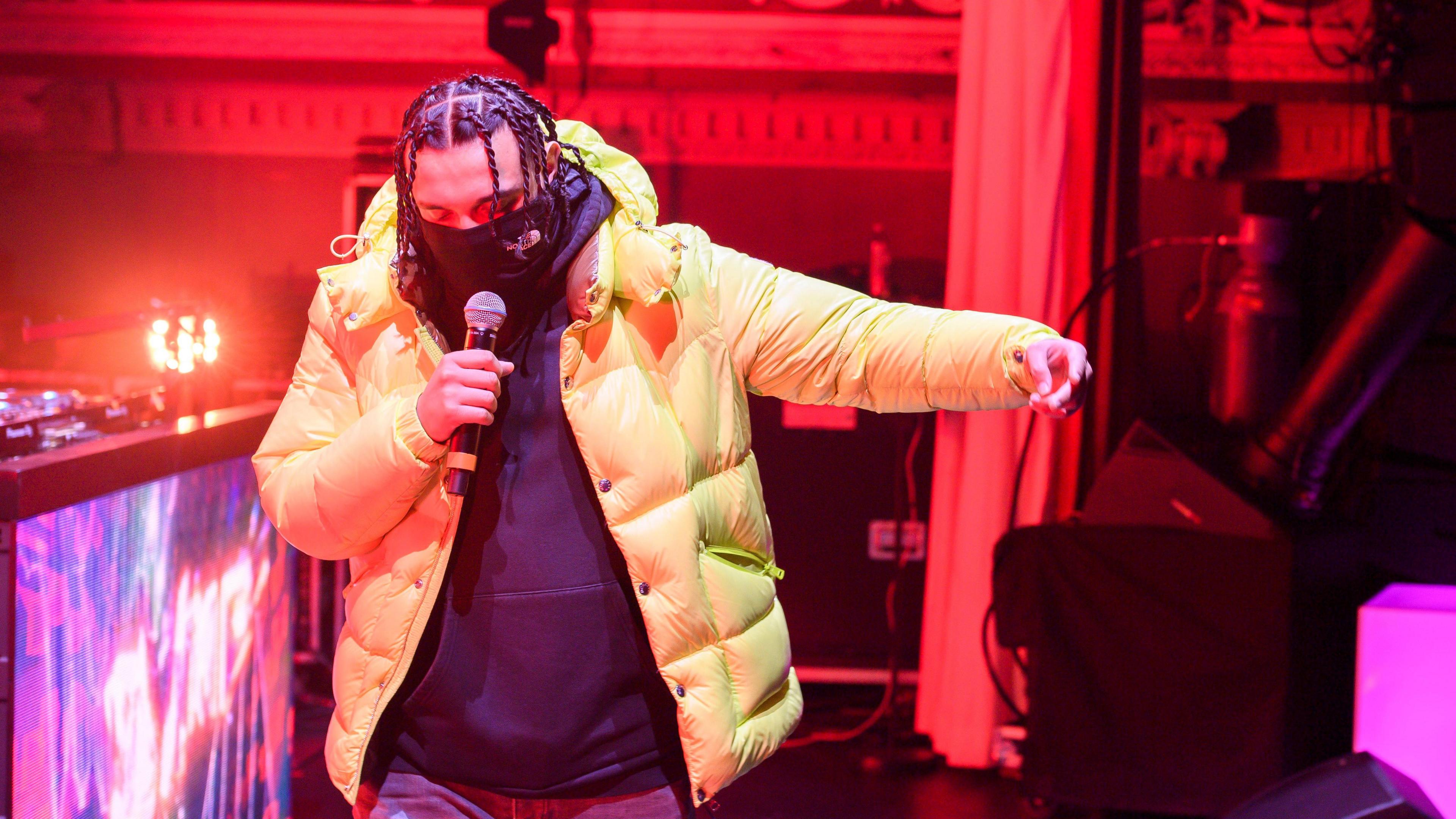 Sliime performing on stage in February 2024. The rapper wears a bright green puffer jacket over a black hoodie and covers the bottom half of his face with a black scarf.  He holds a mic in his right hand, his left outstretched to his side. The staging behind him is lit red and purple. 