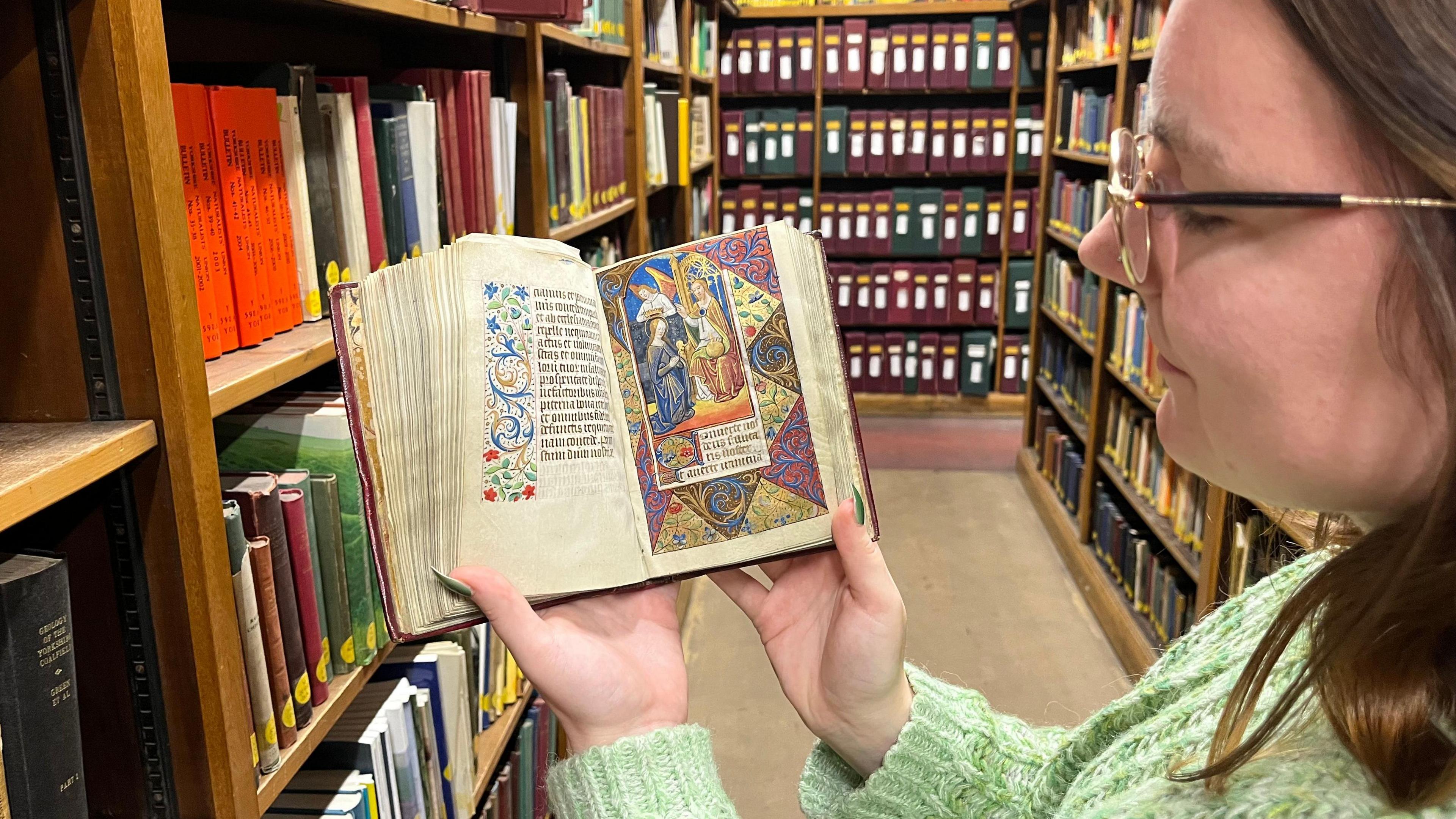 A woman with glasses and long brown hair wearing a green jumper holds the Book of Hours while standing in a library.