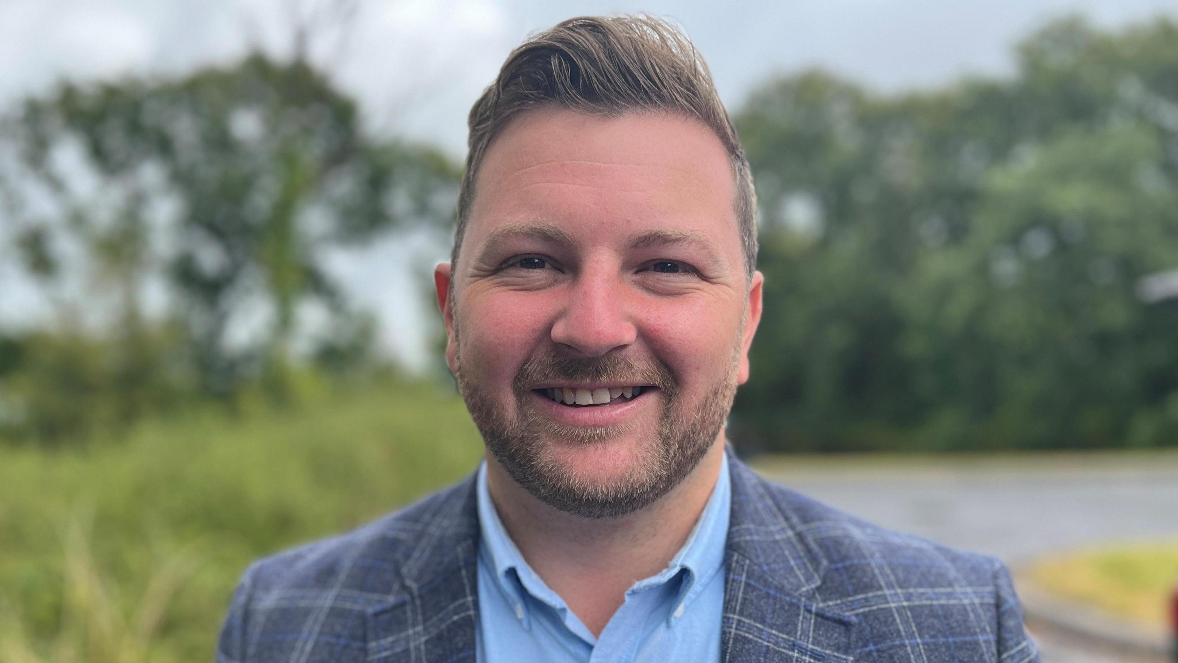Sam Kurtz smiles at the camera. He has brown eyes and short dark hair and a beard, and is wearing a grey plaid blazer and light blue shirt. Trees and grass can be seen blurred behind him.